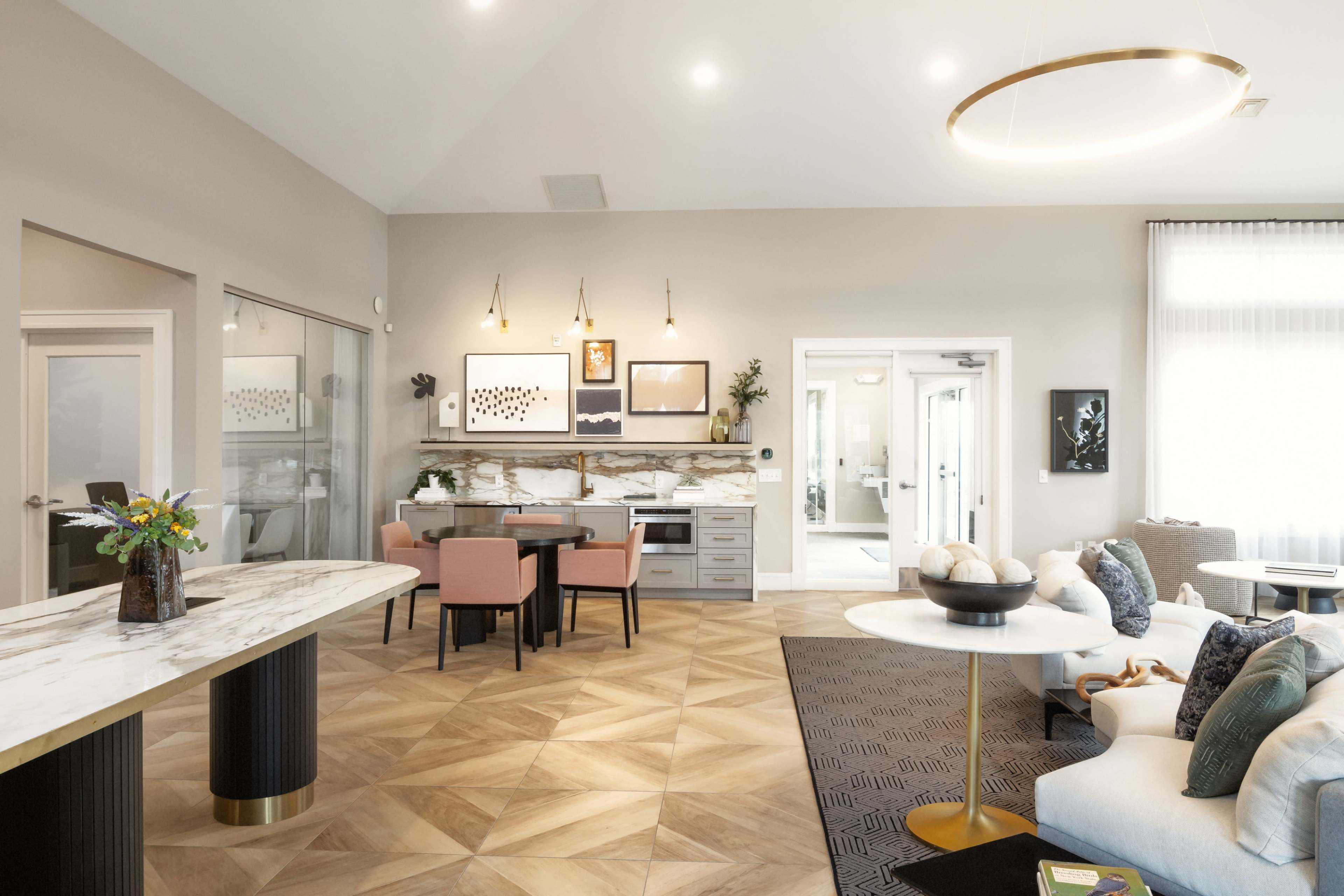The image shows a modern lounge area with a dining table, stylish seating, and decorative wall art, featuring a mix of light-colored walls and patterned flooring.