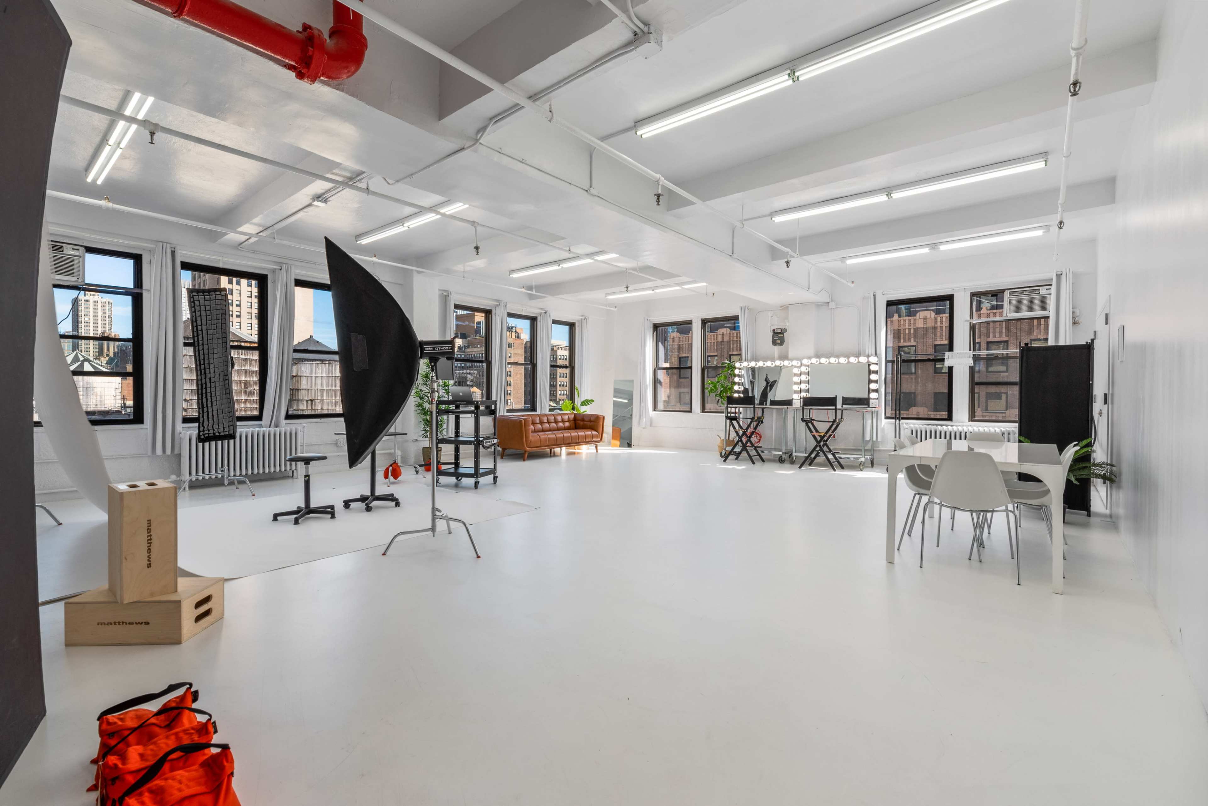 A spacious, bright studio with white walls and floor, equipped with photography lighting, backdrop stands, and furniture.