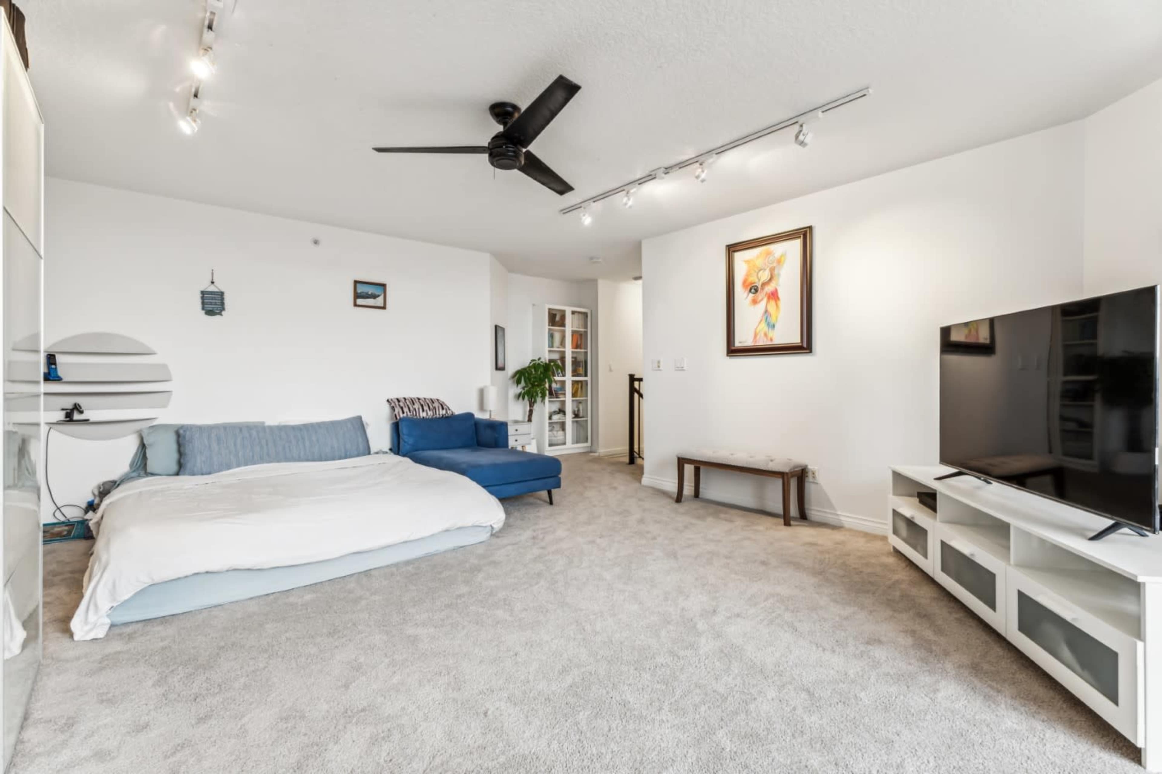 The image shows a modern living space with a blue sofa, a white bed spread on the floor, a television on a white unit, and a ceiling fan.
