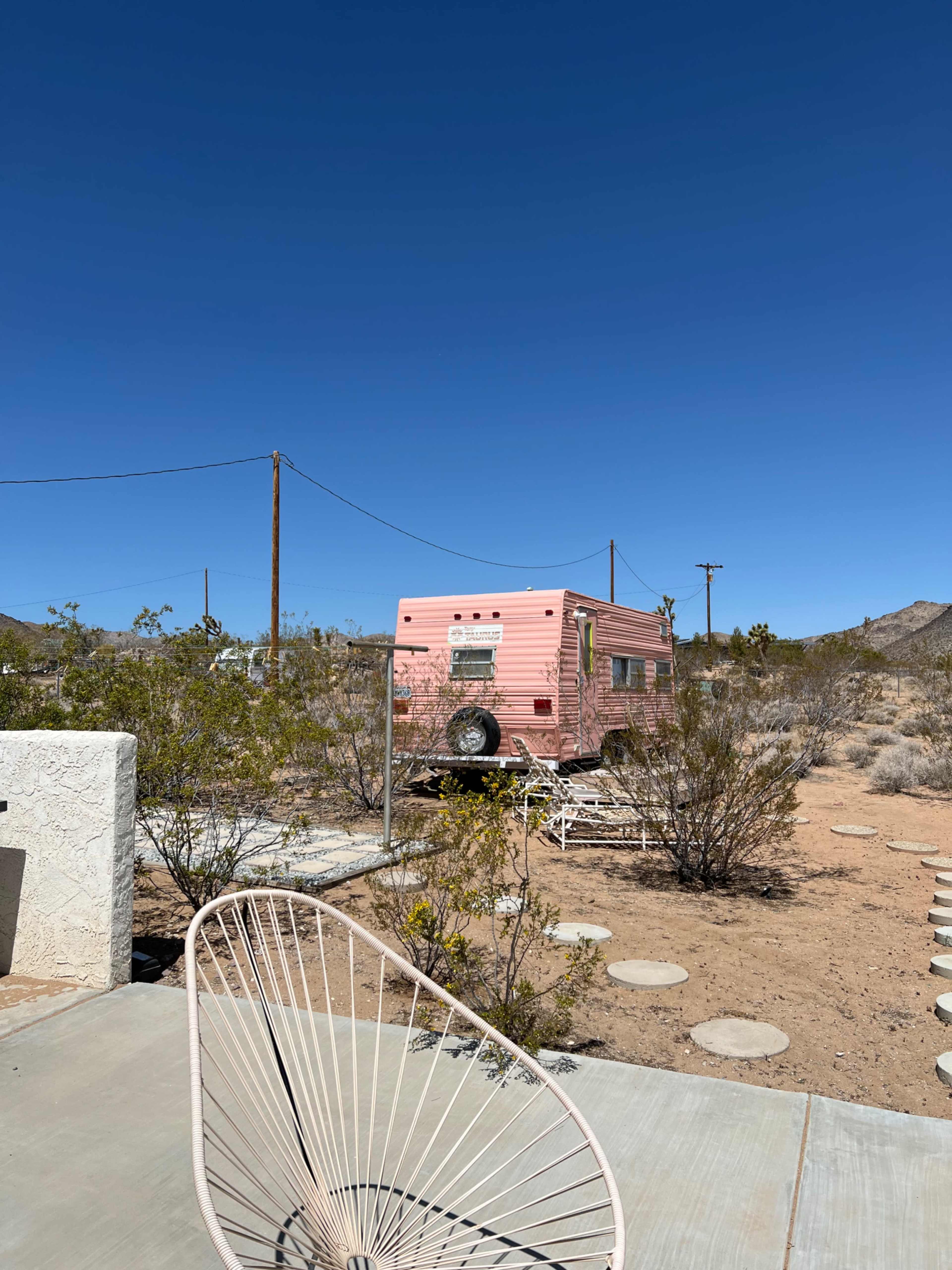 A pink vintage camper is parked on a lot surrounded by sparse desert vegetation under a clear blue sky.