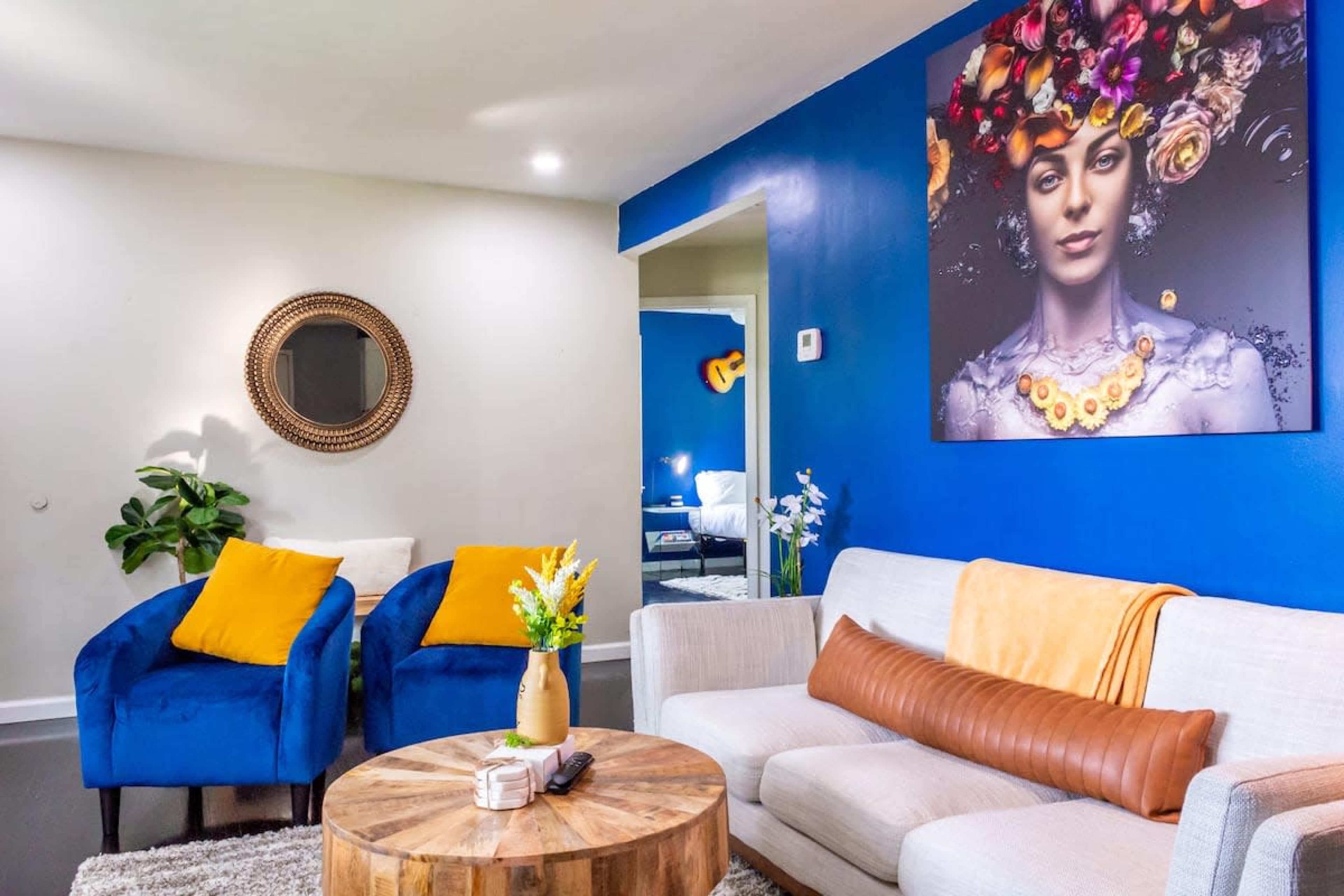 A brightly colored living room features a circular wooden coffee table, a gray sofa with an orange throw, and blue chairs with yellow pillows, alongside a large portrait on the wall.