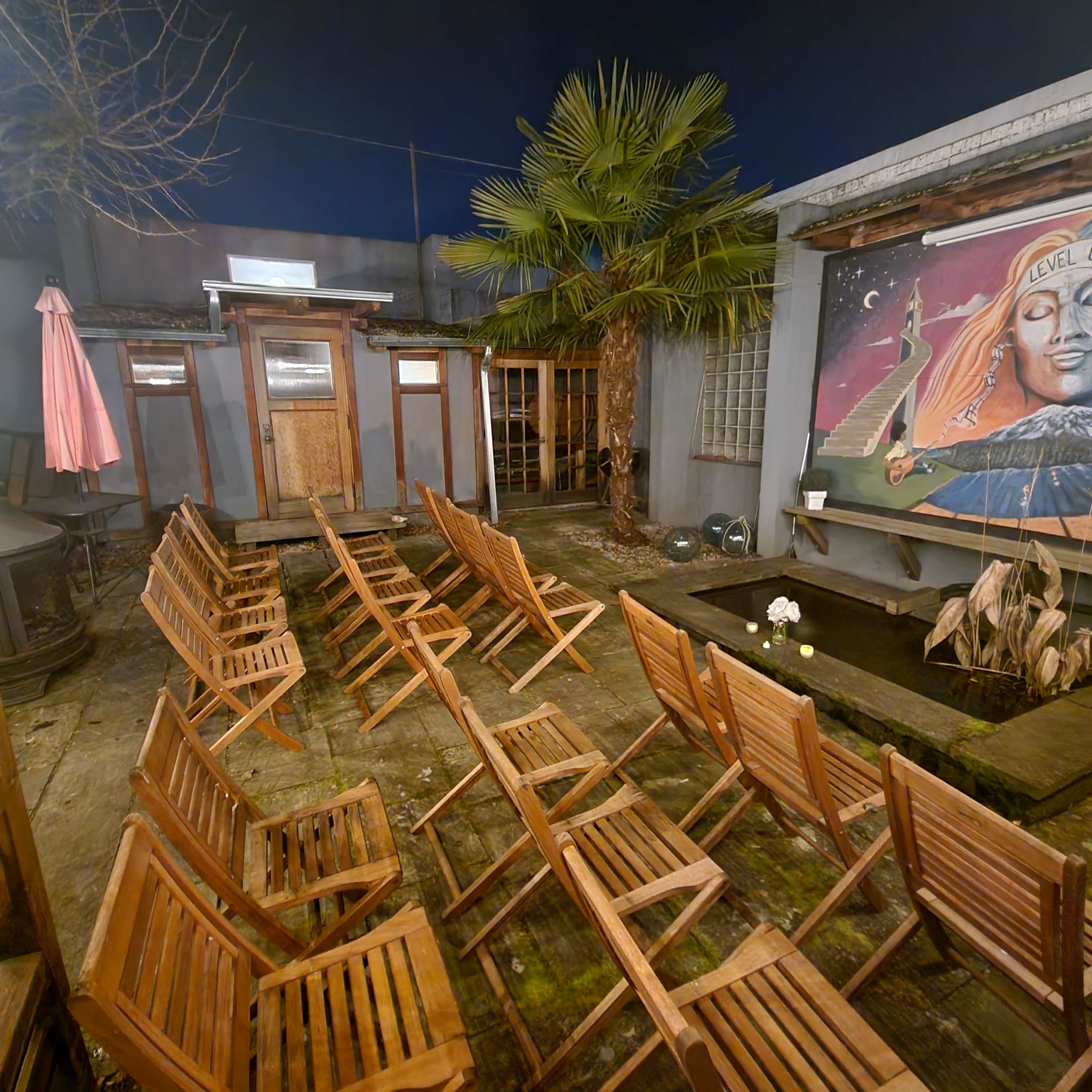 An outdoor seating area with wooden chairs arranged in rows facing a small pond and a mural featuring a figure with a palm tree in the background.