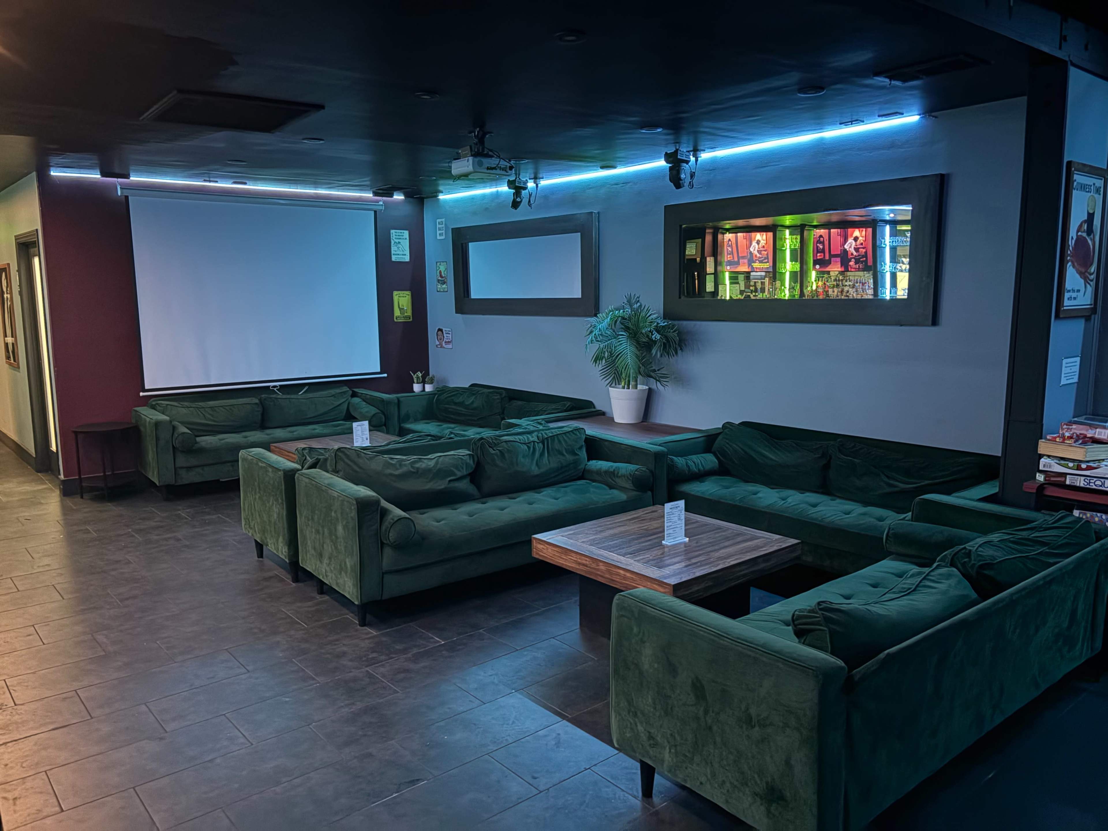 The image shows a lounge area with green sofas arranged around a wooden table, illuminated by blue LED lights, and featuring a large screen on one wall.