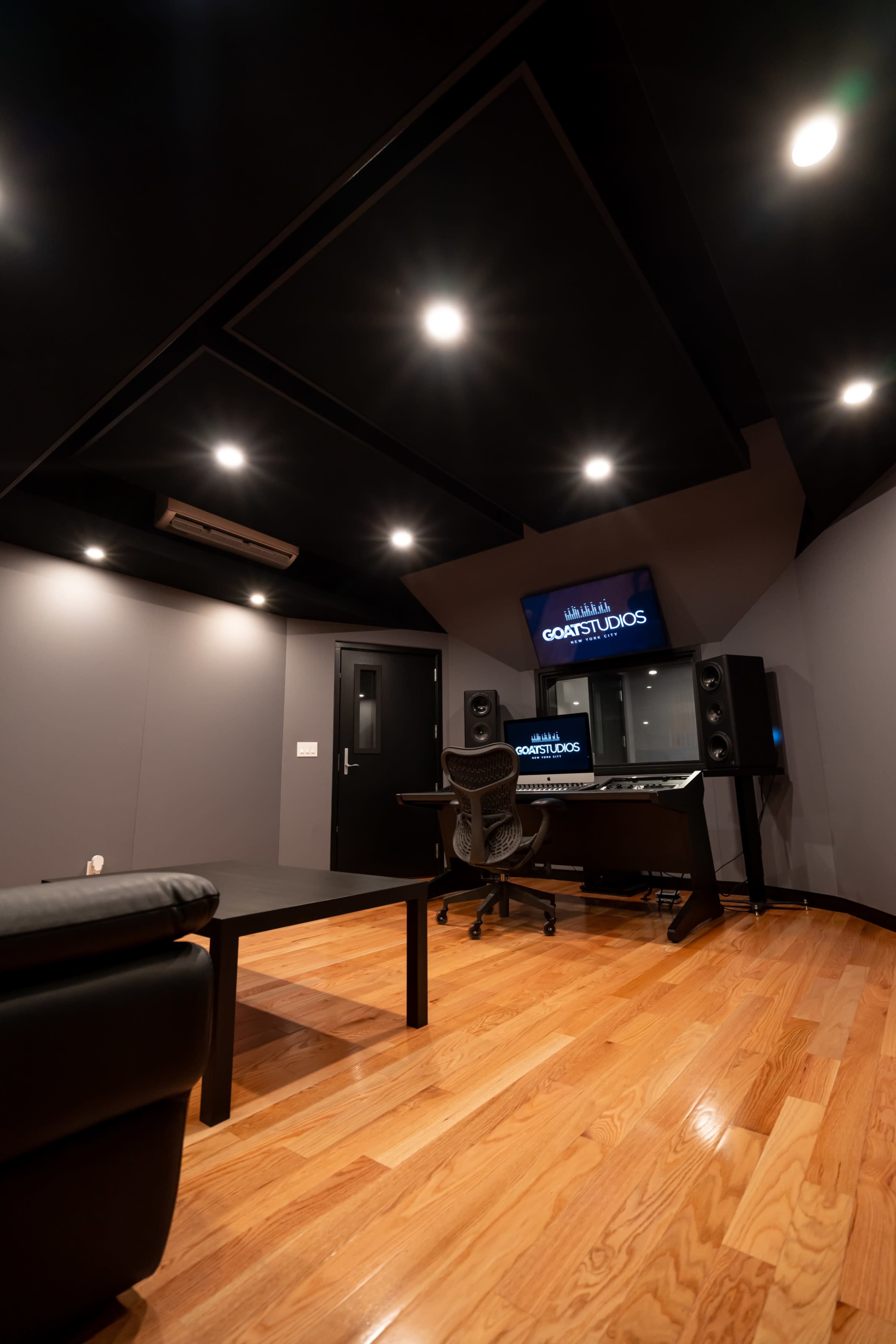 A modern recording studio with a desk, speakers, and a large screen displaying "GOAT STUDIOS" on the wall.