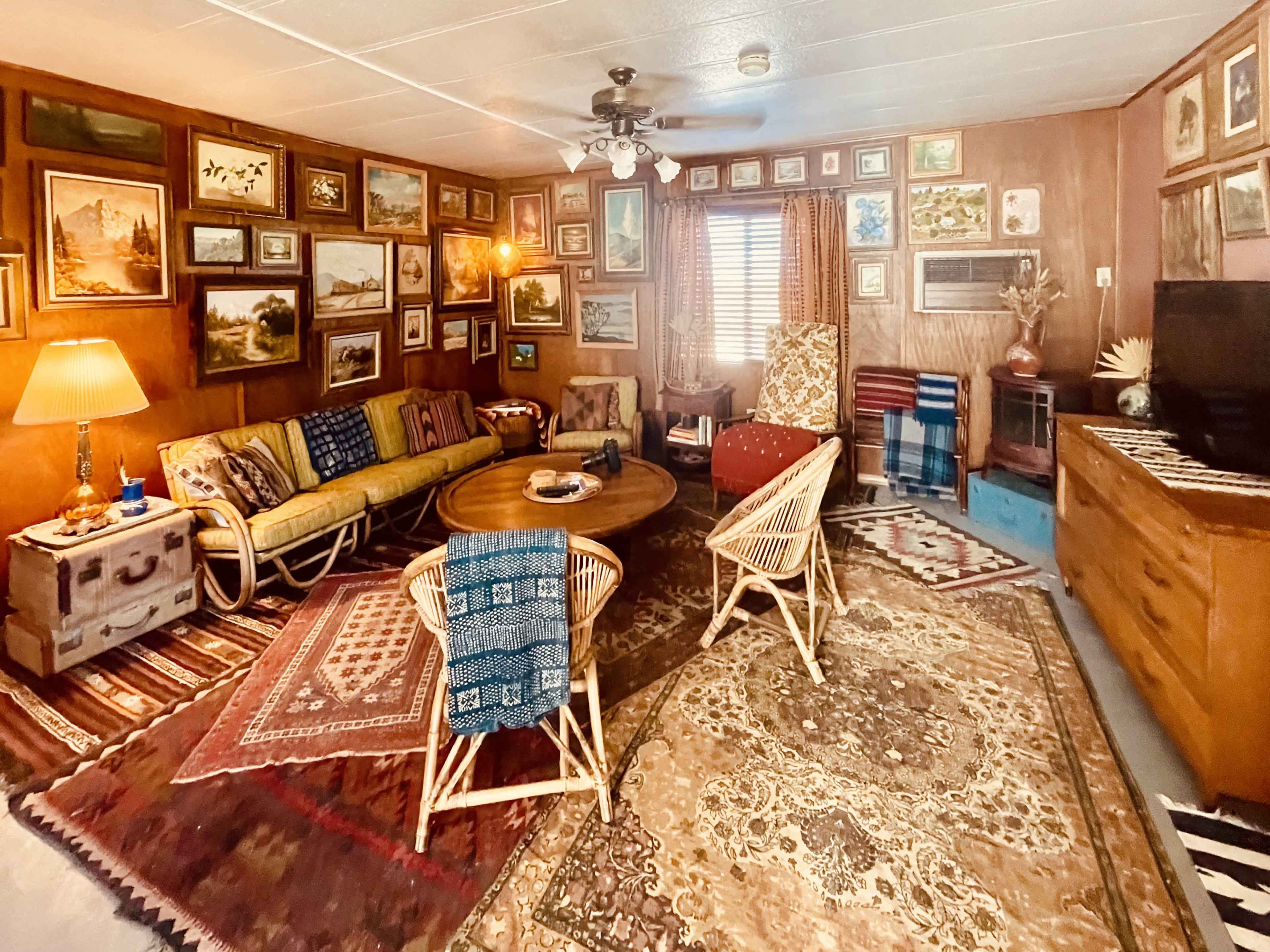 The room features a collection of framed artwork on wooden walls, with a central round table surrounded by chairs and a mix of rugs covering the floor.