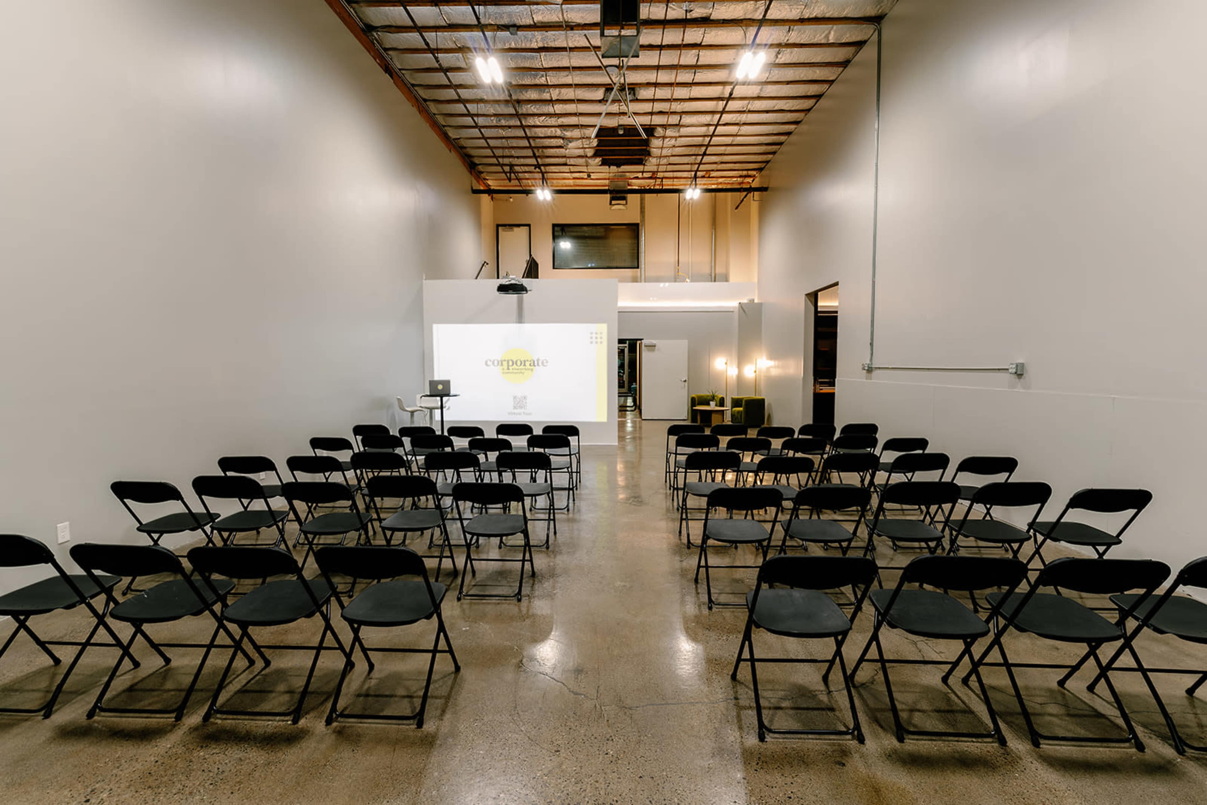 A spacious event room features rows of black folding chairs set up for a presentation, with a projector screen at the front.