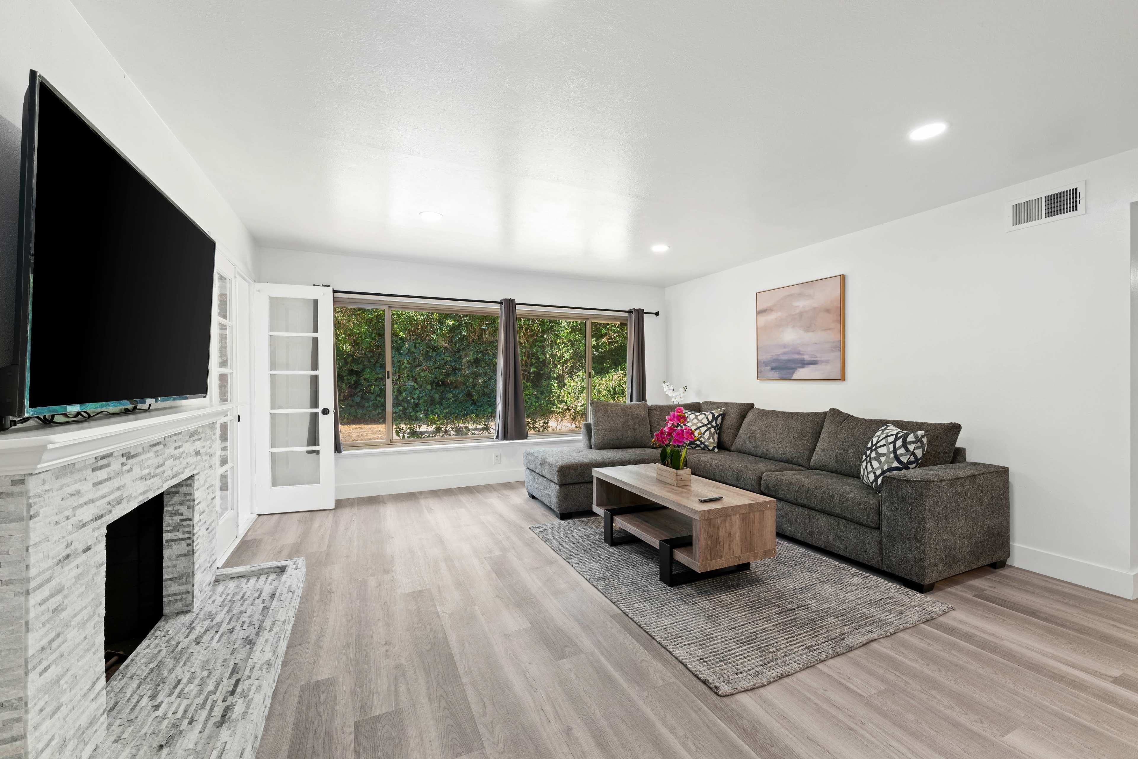 A modern living room with a gray sectional sofa, a wooden coffee table, and a large TV mounted on the wall, alongside a window offering a view of greenery outside.