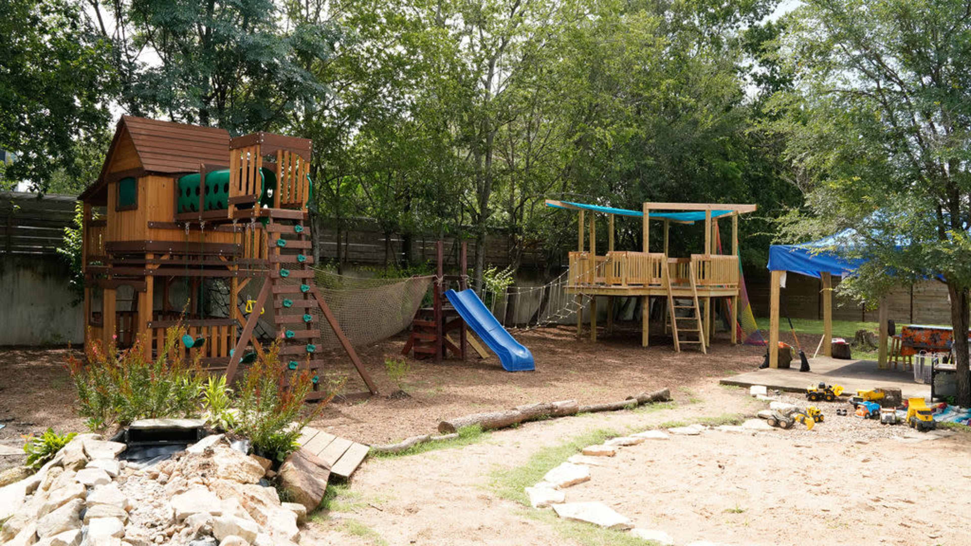 The image shows a playground with a wooden play structure featuring a slide and climbing features, along with a separate elevated wooden platform and a sandy play area.