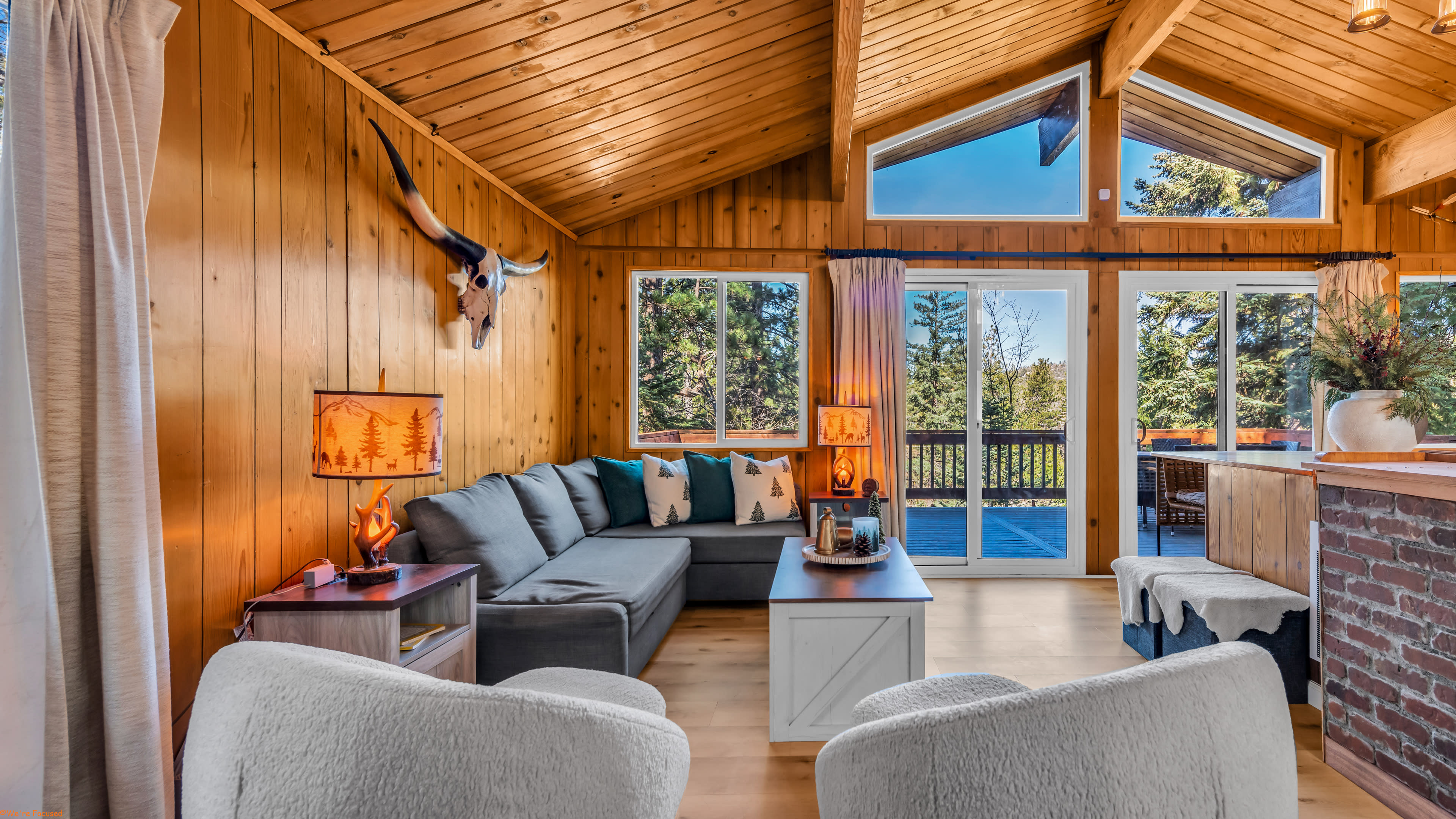 The image shows a cozy living room with wooden walls, a large gray sectional sofa, and large windows that provide a view of the outdoors.