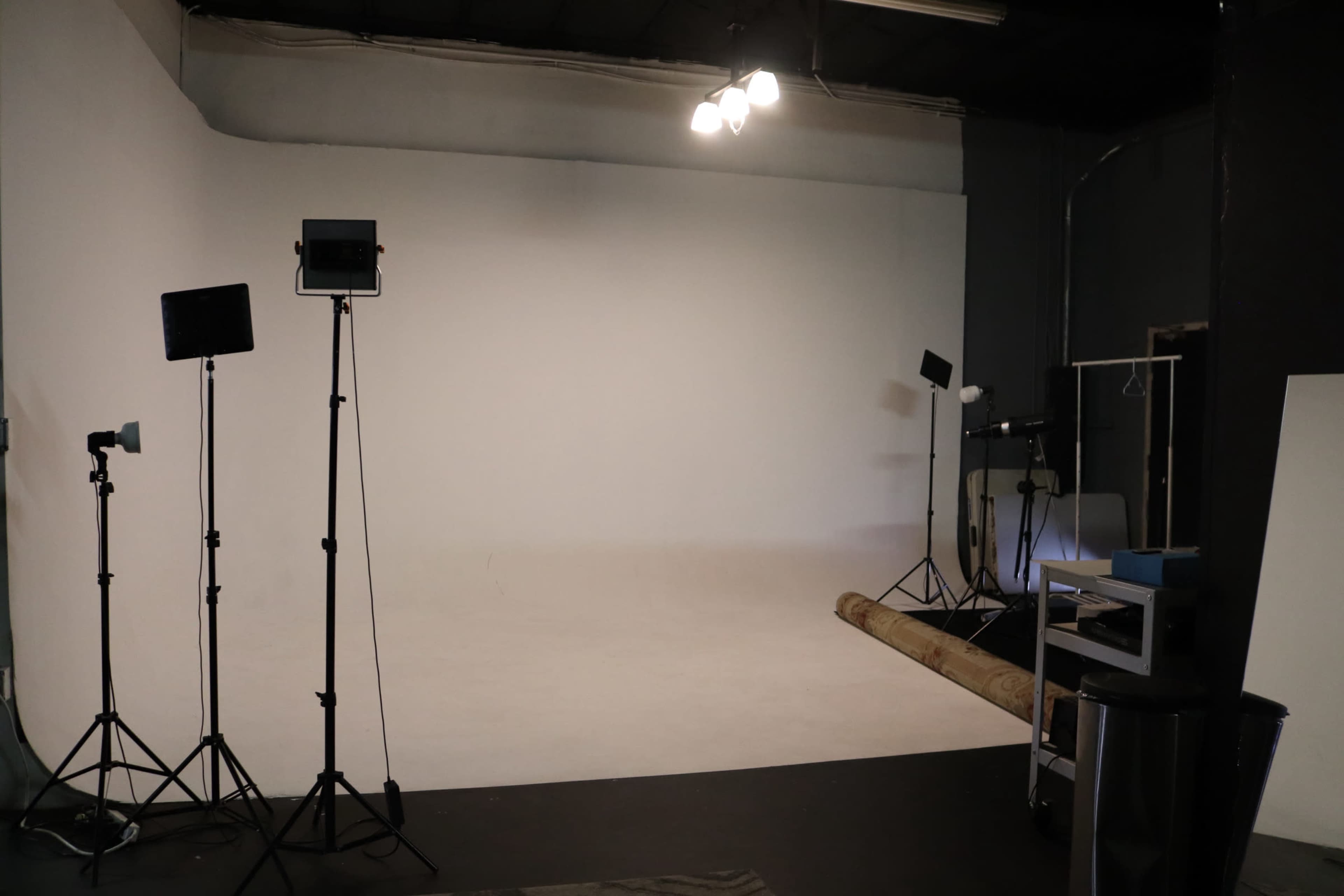 The image shows a photography studio with a blank backdrop, tripod-mounted lights, and a roll of seamless paper on the floor.