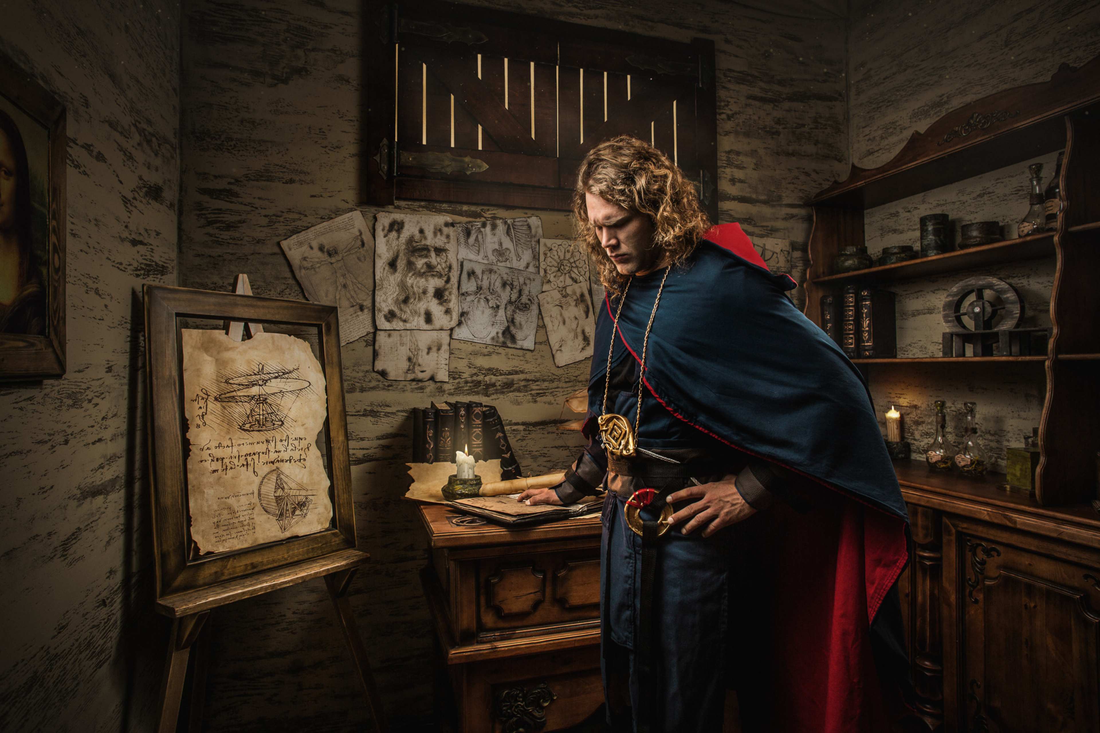 A man in a long, dark cloak examines a parchment on an easel in a dimly lit room filled with old furniture and various artifacts.