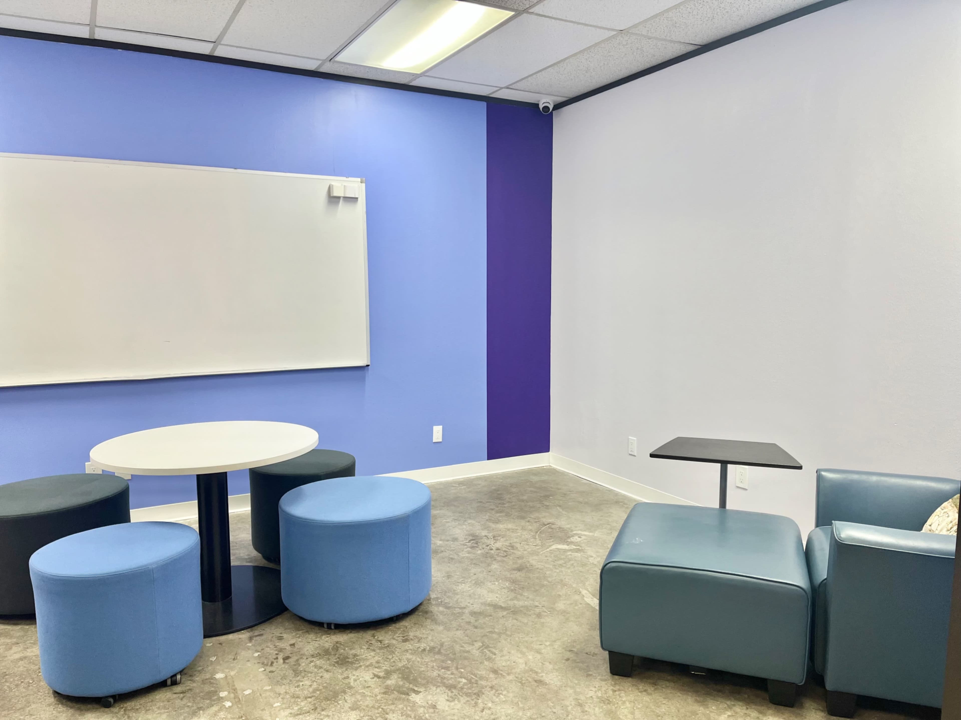 The image shows a small meeting space with a round table surrounded by four blue ottomans and a blue armchair with a small side table.