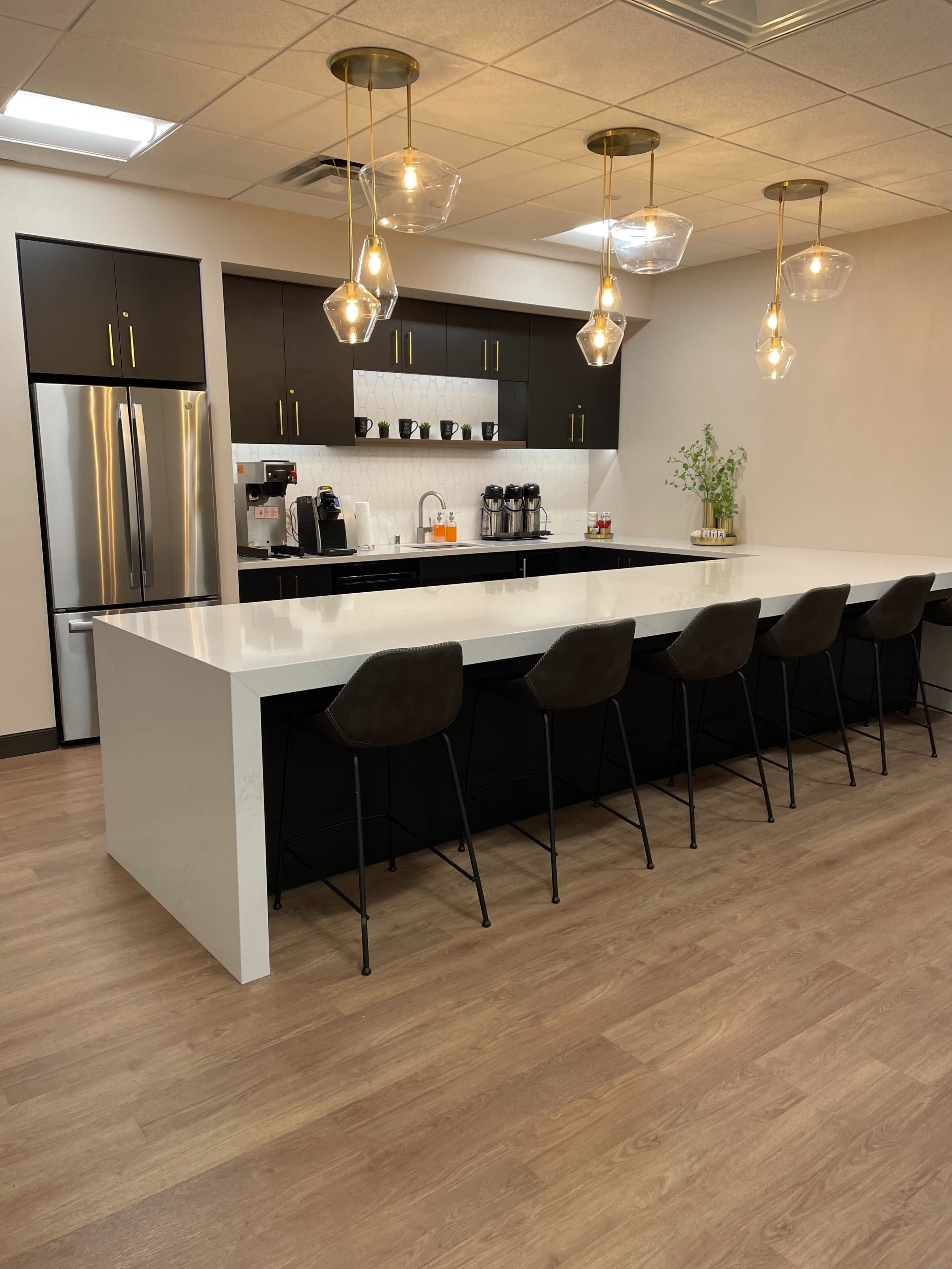 A modern kitchen features a large white island with bar stools, dark cabinets, a stainless steel refrigerator, and pendant lighting.