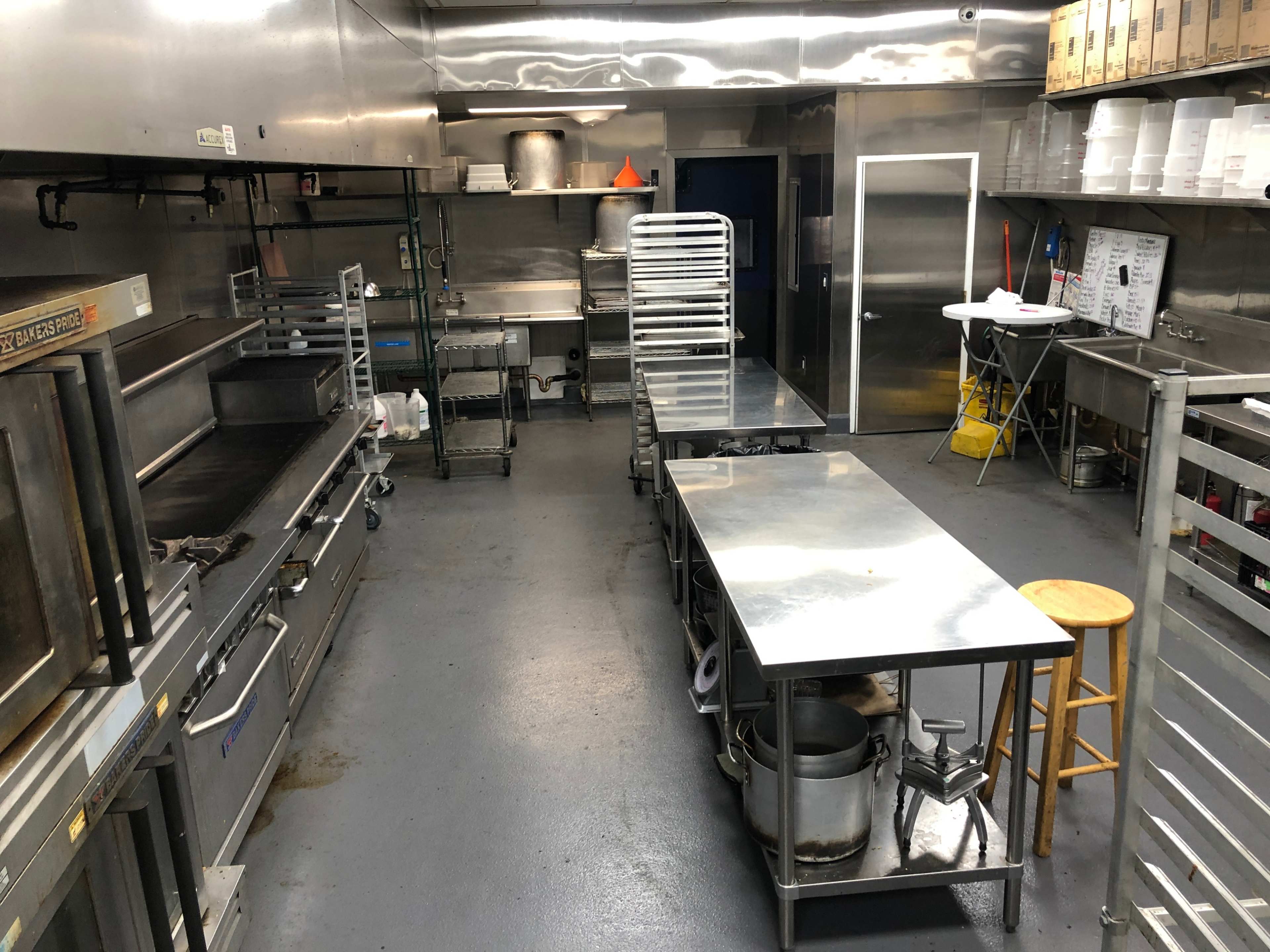 The image shows a stainless steel commercial kitchen with multiple worktables, cooking equipment, and shelves.