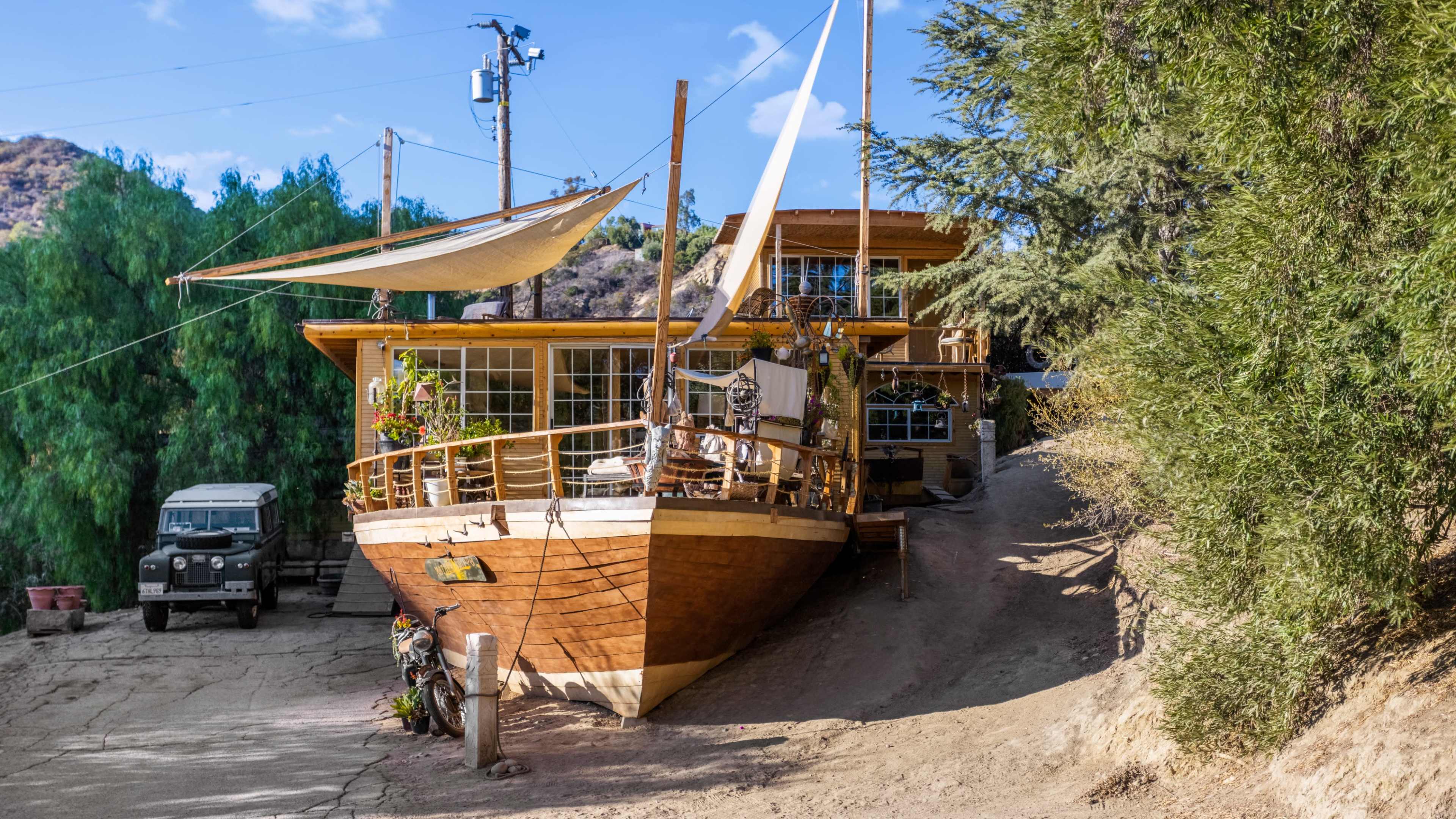 A wooden boat is converted into a house situated on a hillside, with a dirt path leading up to it and a vintage vehicle parked nearby.