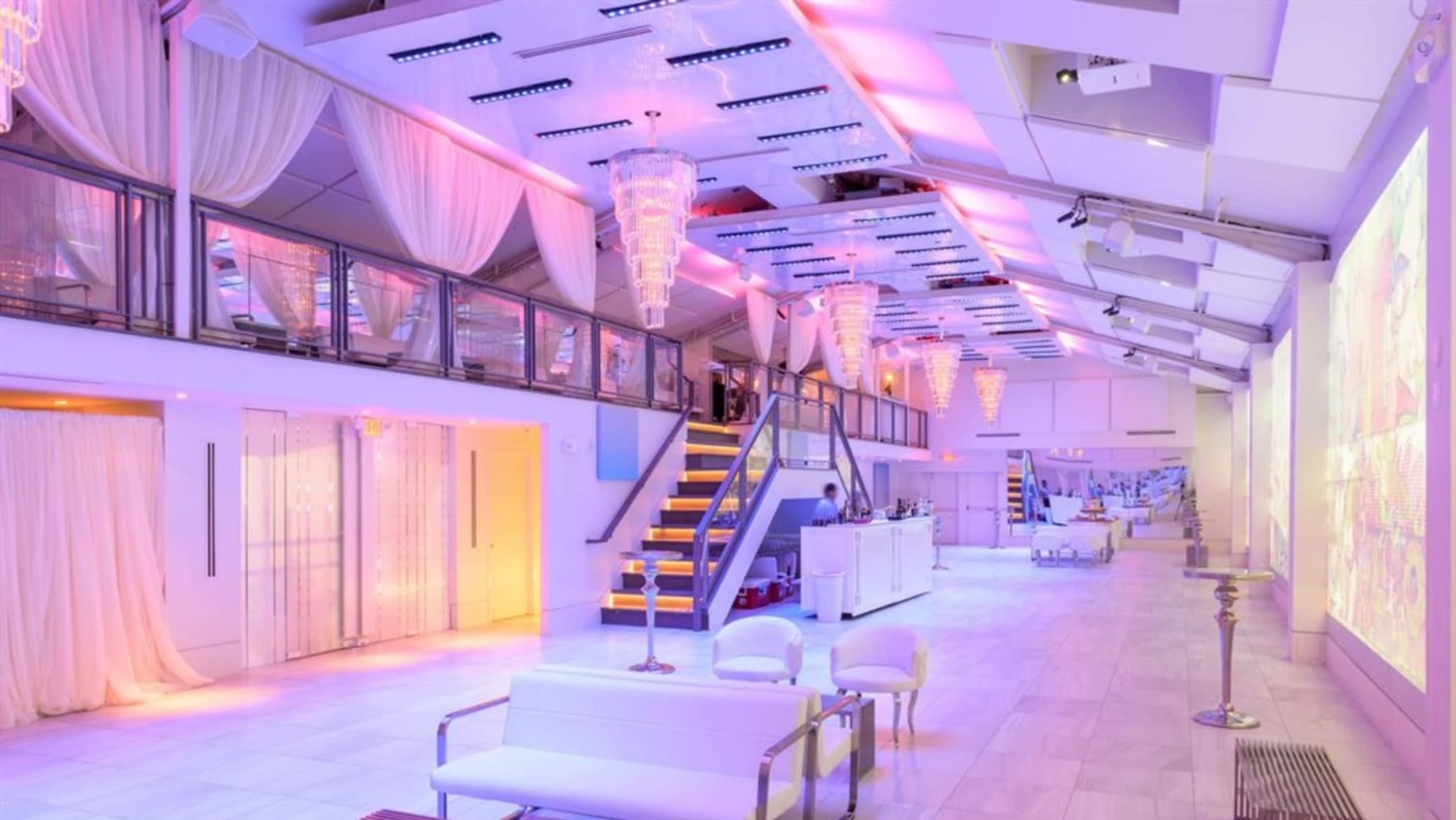 The image shows a brightly lit modern event space featuring white decor, chandeliers, and a staircase leading to an upper level.