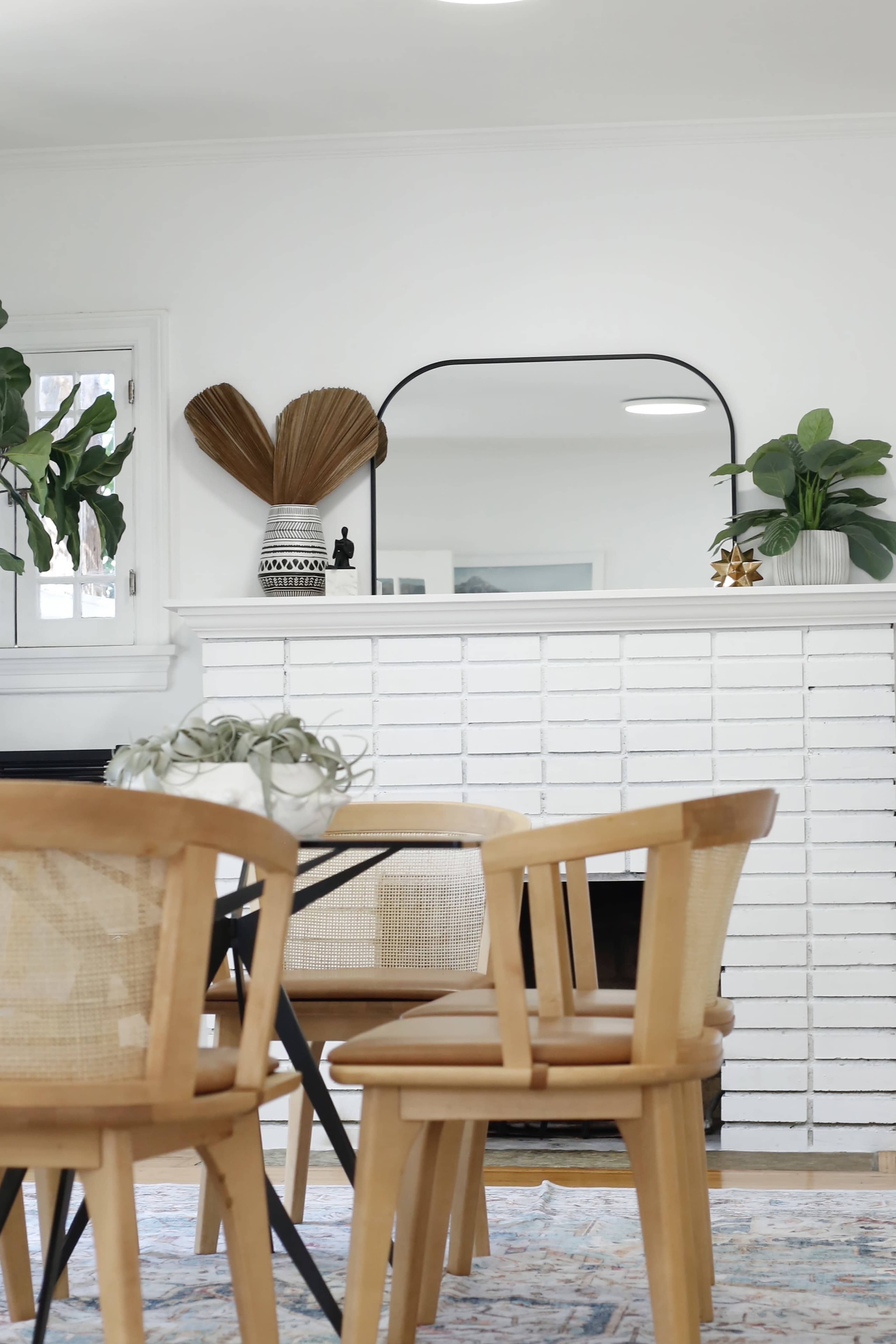 A bright room features a white brick fireplace with plants and decorative items on the mantel, while a wooden table and chairs are arranged in the foreground.