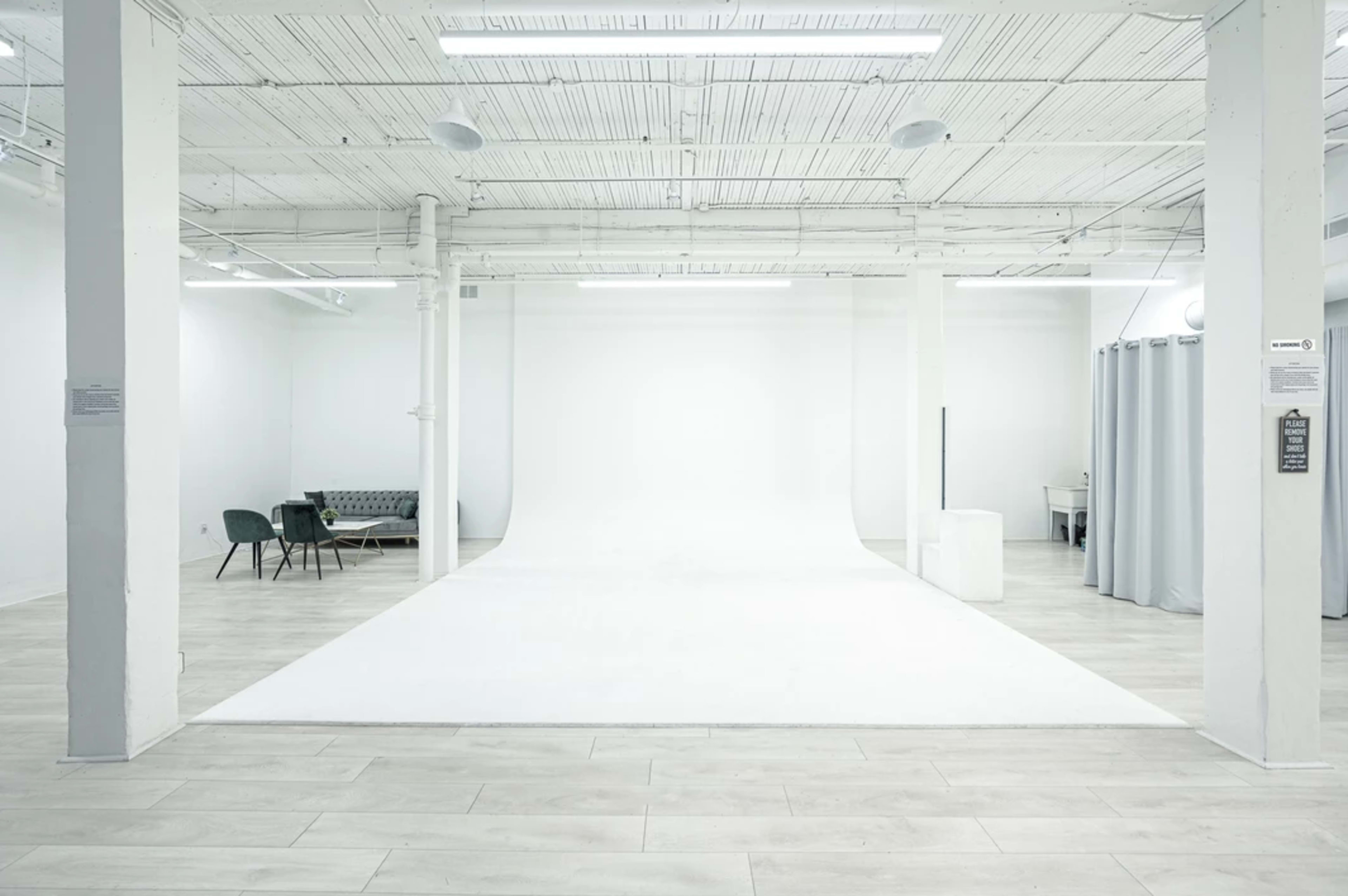 A spacious, modern studio with a white backdrop, wooden flooring, and a seating area consisting of chairs and a couch.