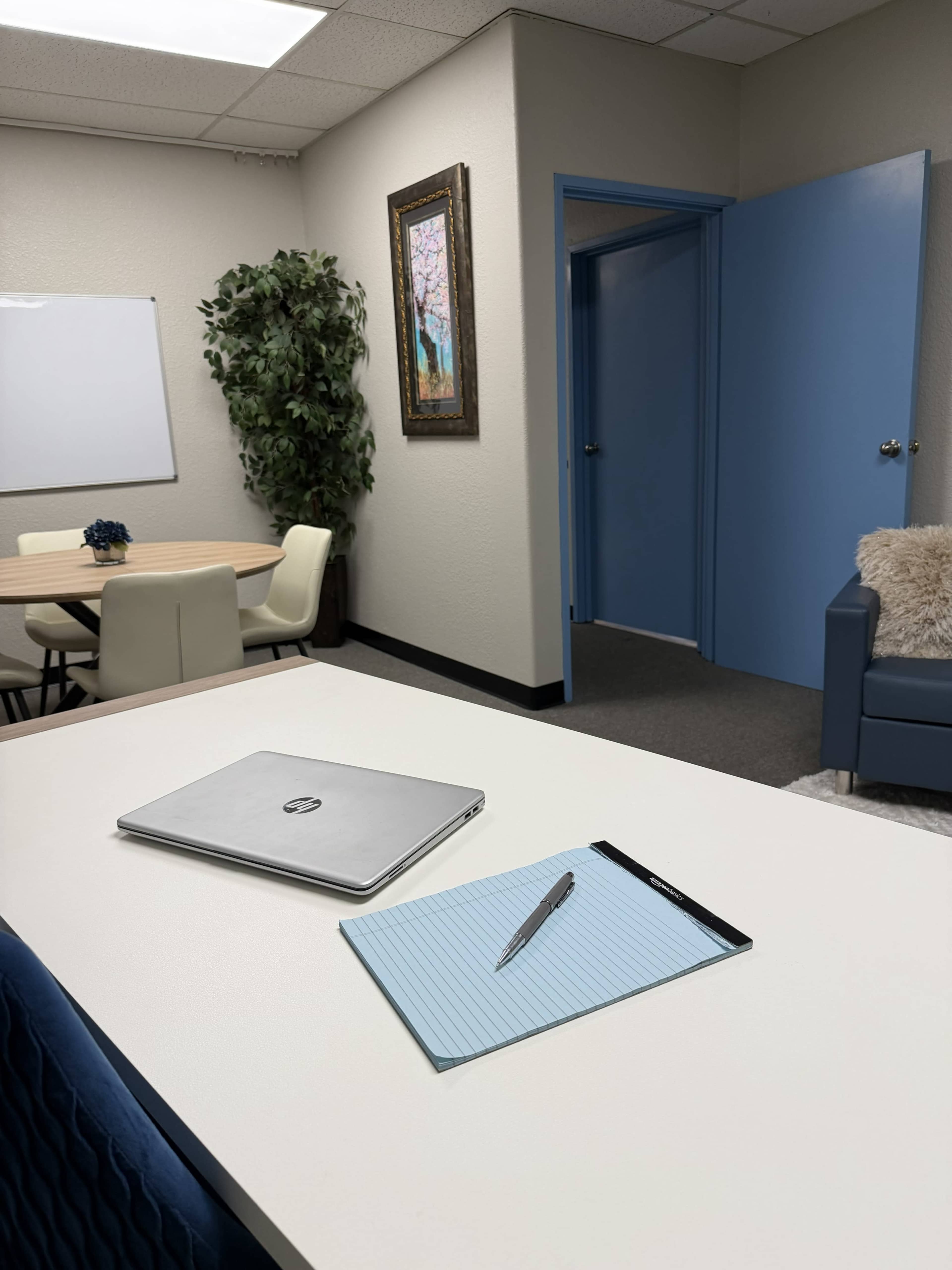 The image shows a modern office space with a desk featuring a laptop, a notepad, and a pen, along with a round table and chairs in the background.