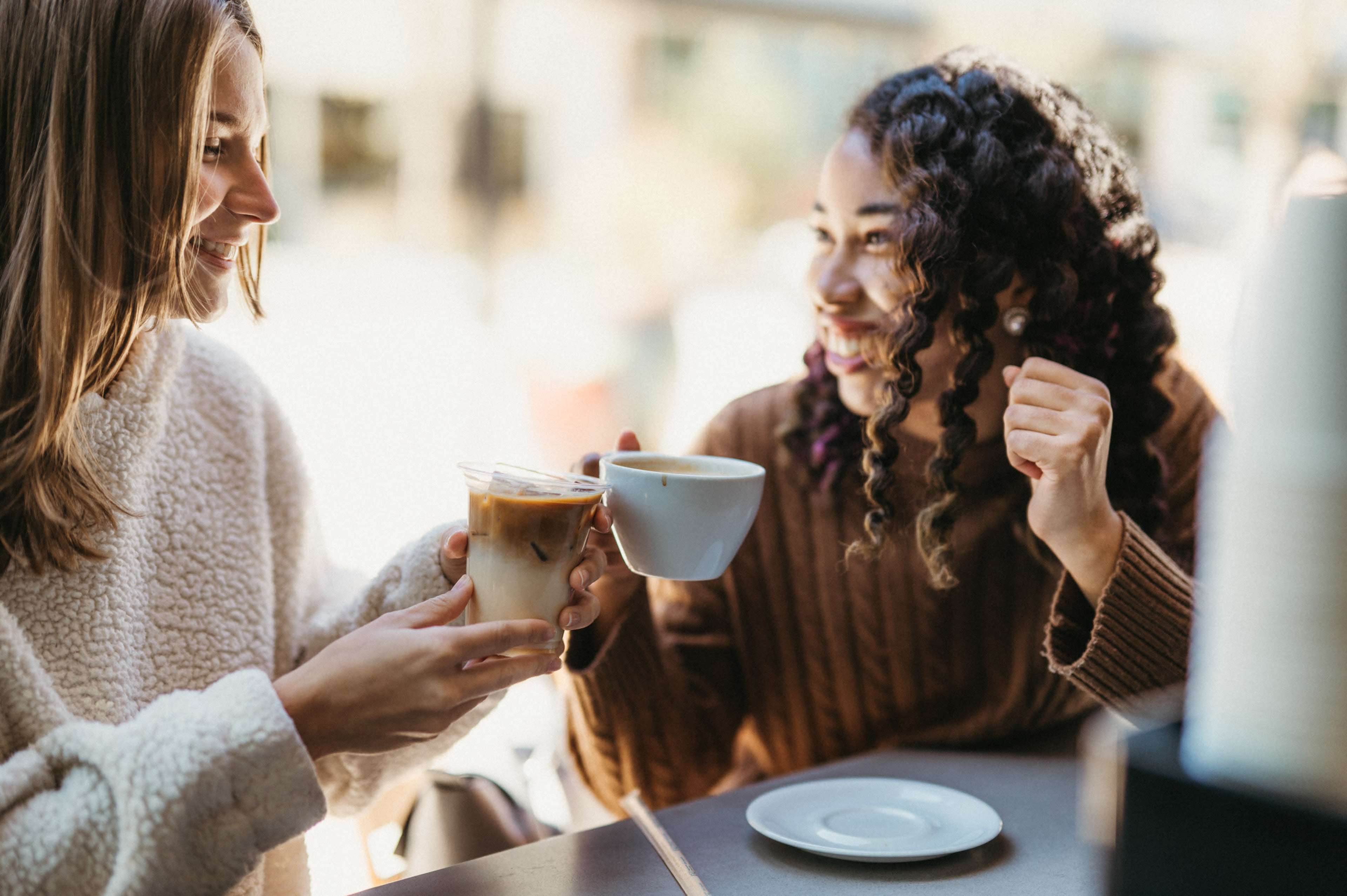 Two women sit at a table and toast their drinks in a bright cafe.