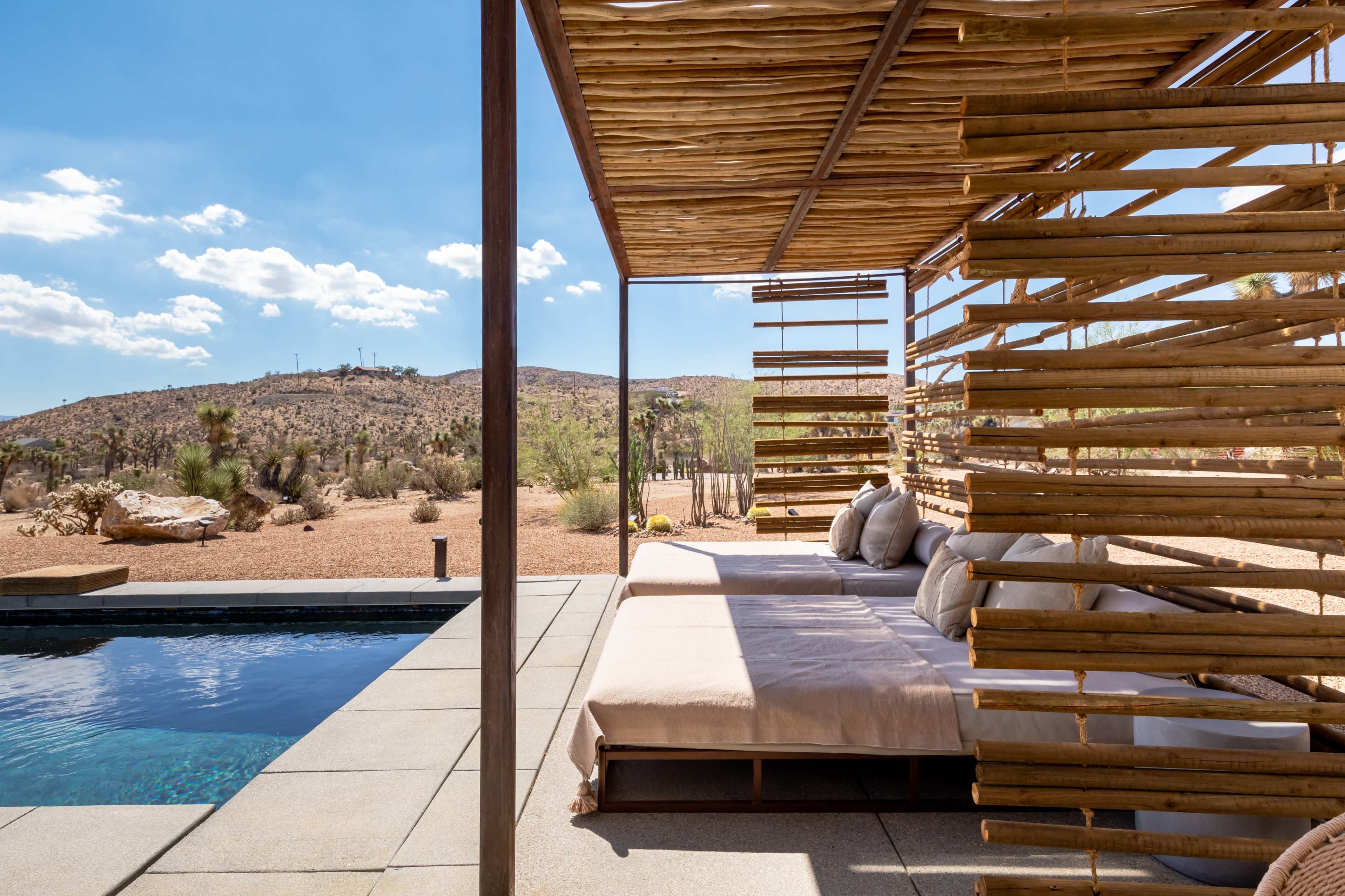 A desert patio features a wooden structure with lounge seating beside a swimming pool and a view of the surrounding landscape.
