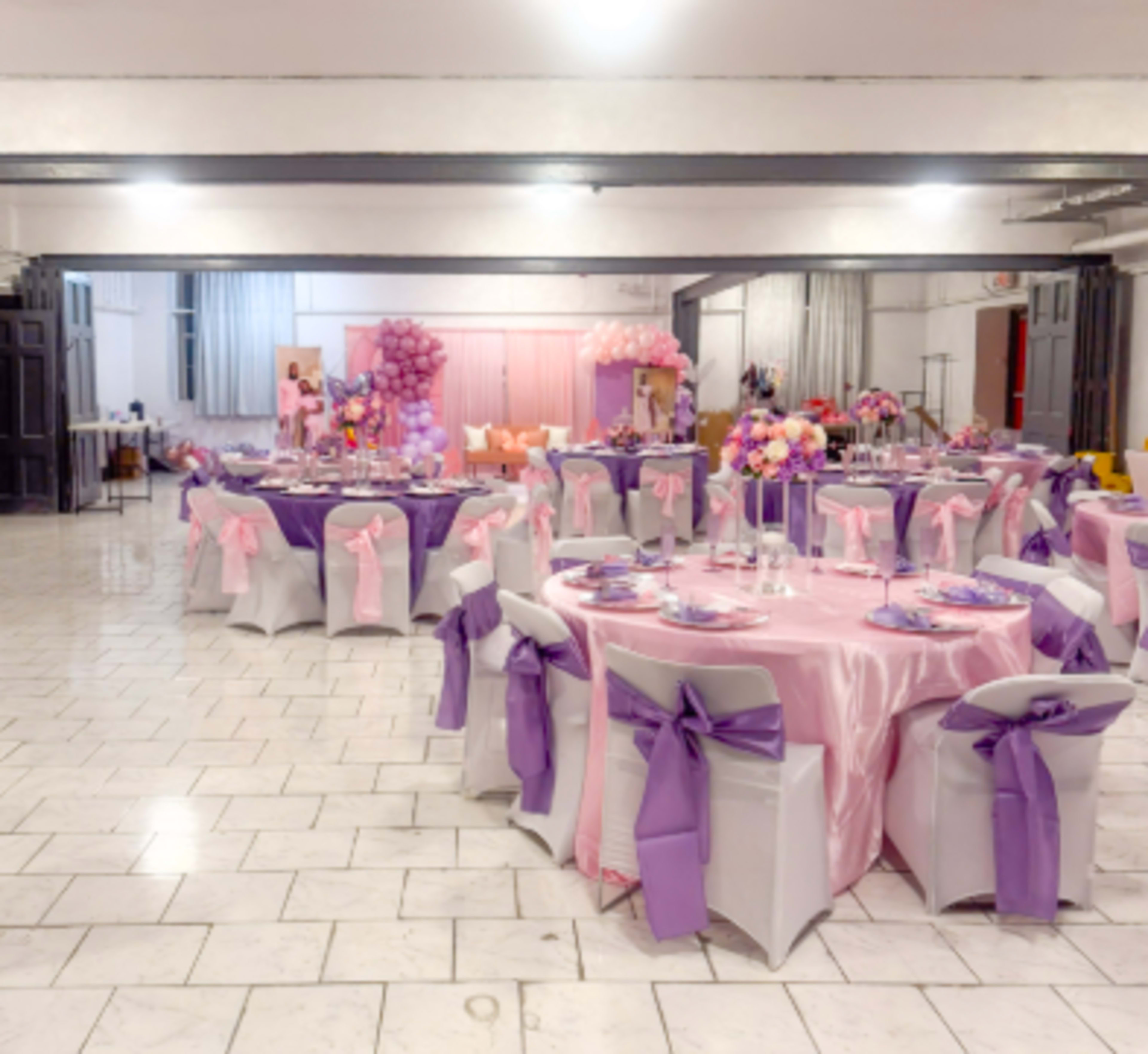The image shows a banquet hall decorated for an event, featuring tables covered in pink and purple tablecloths with matching chair sashes.