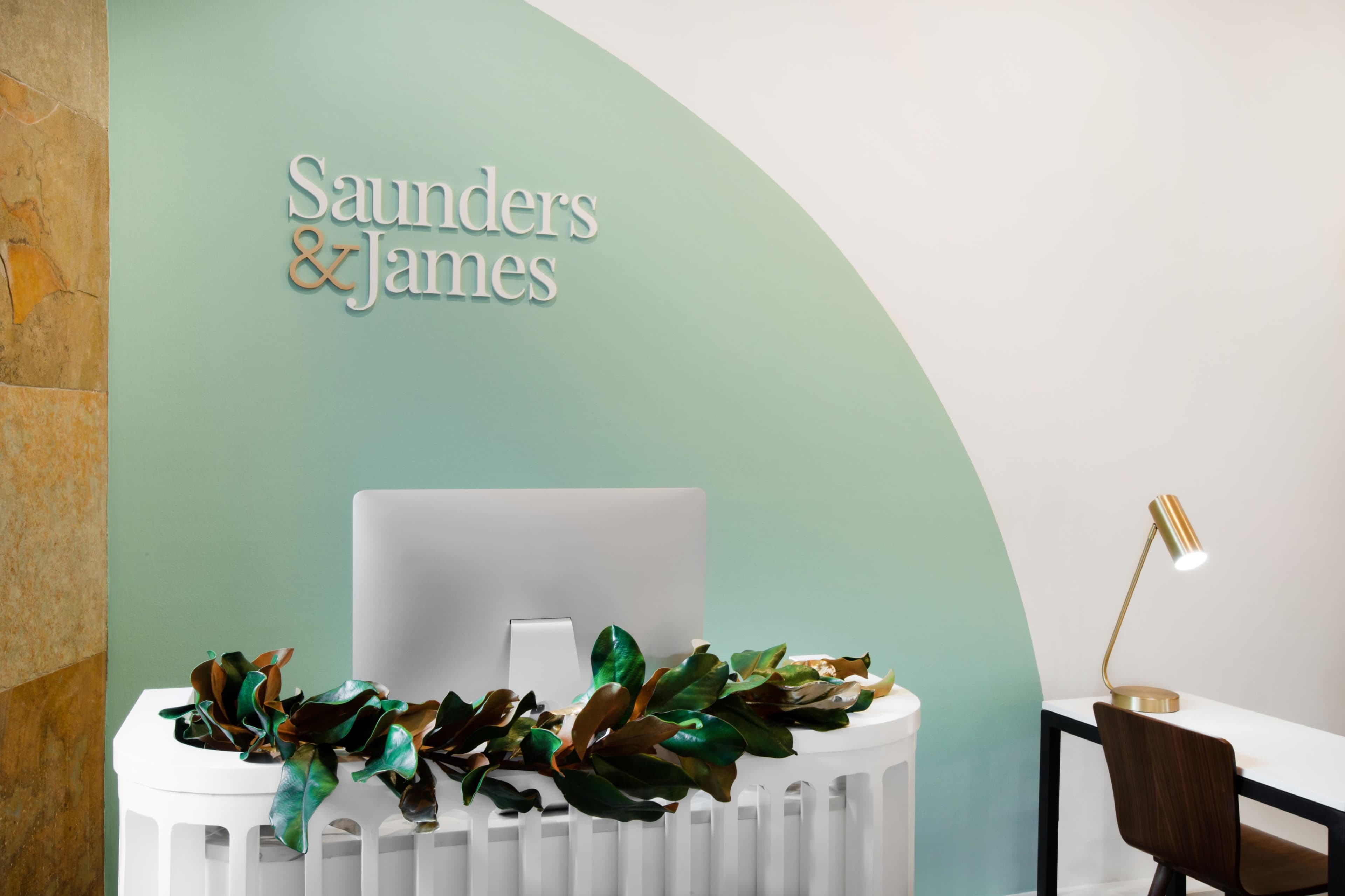 The image shows a reception area with a white desk adorned with a leafy garland, a computer monitor, a desk lamp, and a minimalist chair, next to a wall featuring the name "Saunders & James."