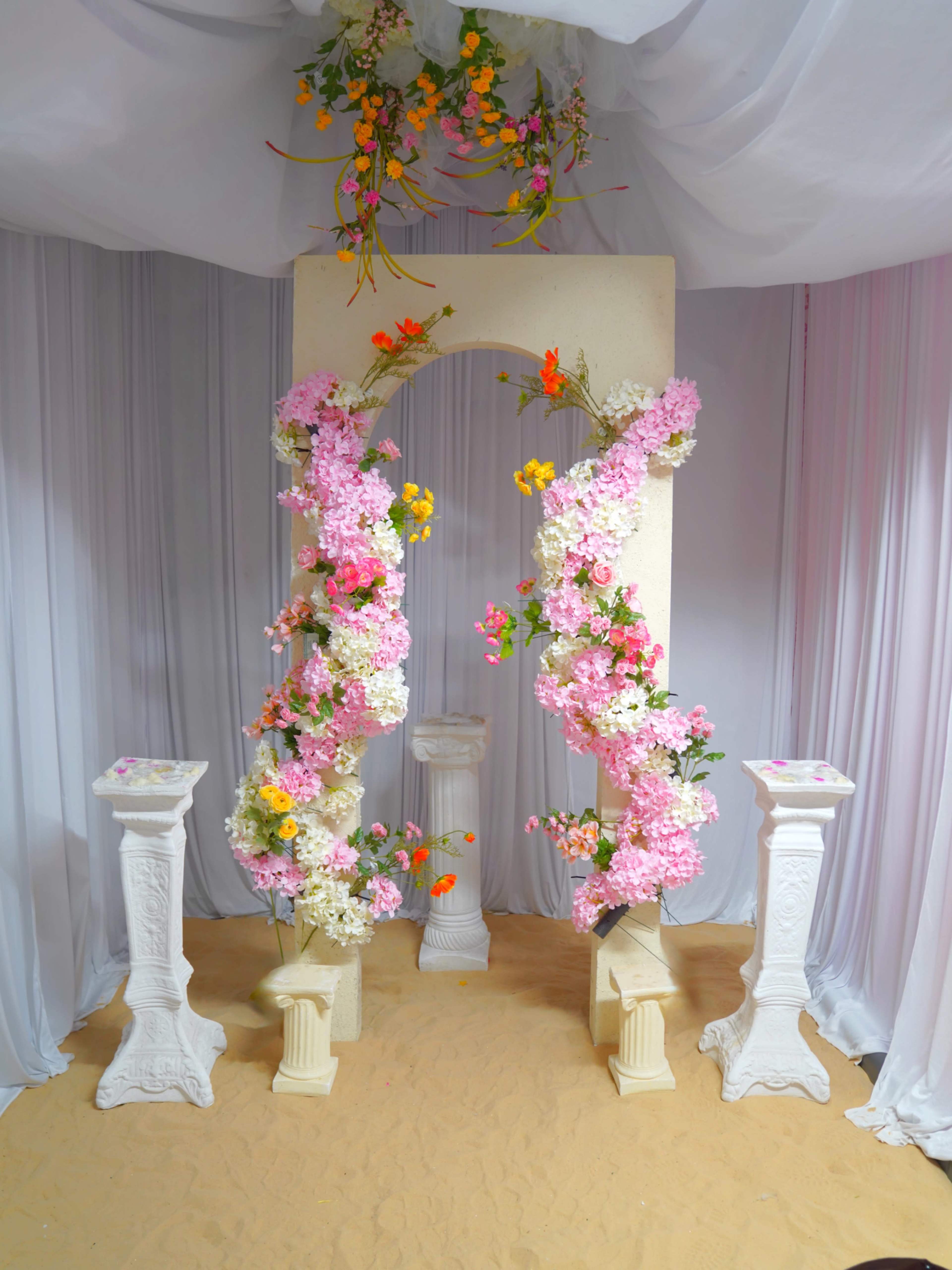 The image shows a decorated wedding arch with floral arrangements in shades of pink and orange, flanked by two white pedestals, all set against a backdrop of draped fabric.