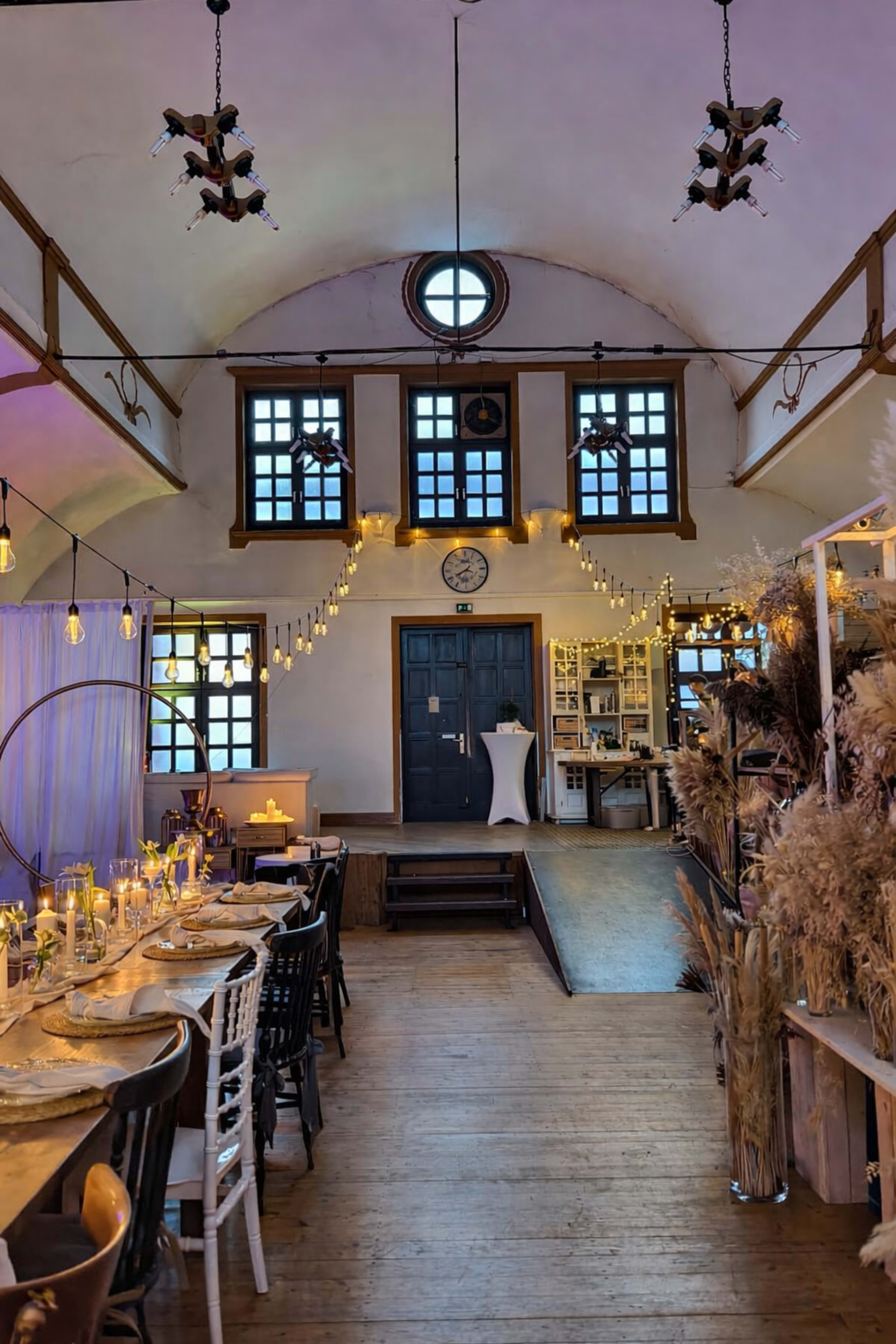 The image shows a warmly lit event space with wooden flooring, rows of tables set for dining, and decorative elements like string lights and pampas grass.