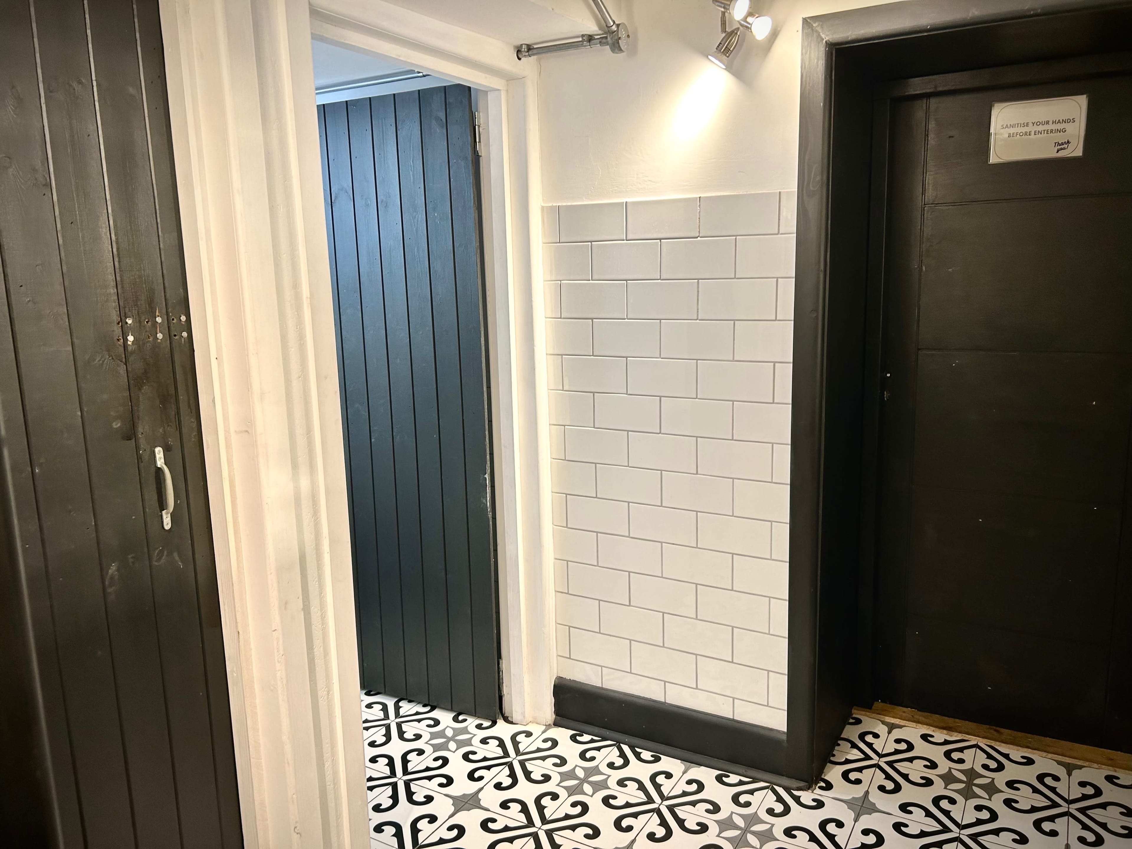 The image shows a hallway with black wooden doors, a section of white tiled wall, and patterned black and white floor tiles.