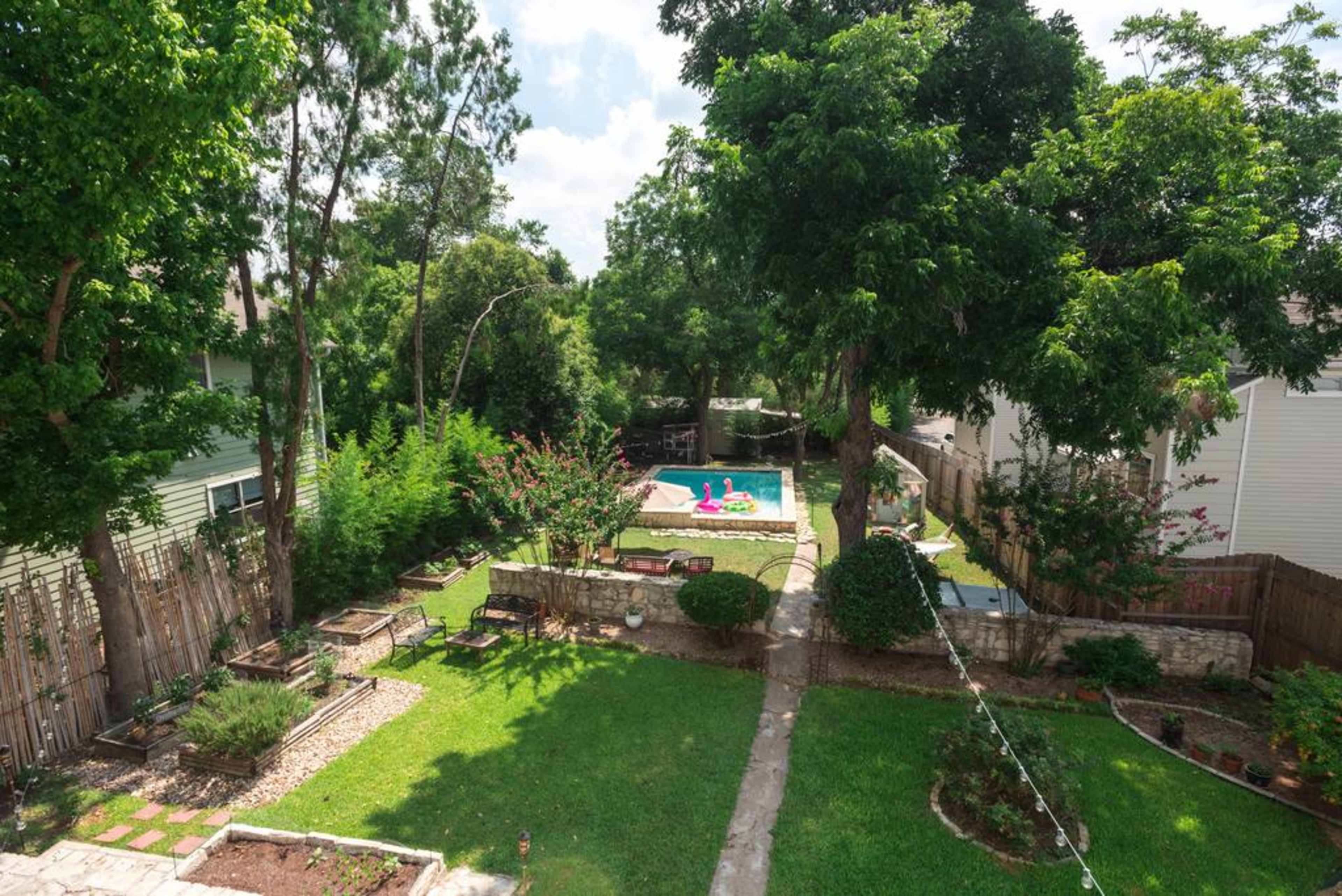 A grassy backyard with a stone path leading to a swimming pool surrounded by trees and colorful pool floats.
