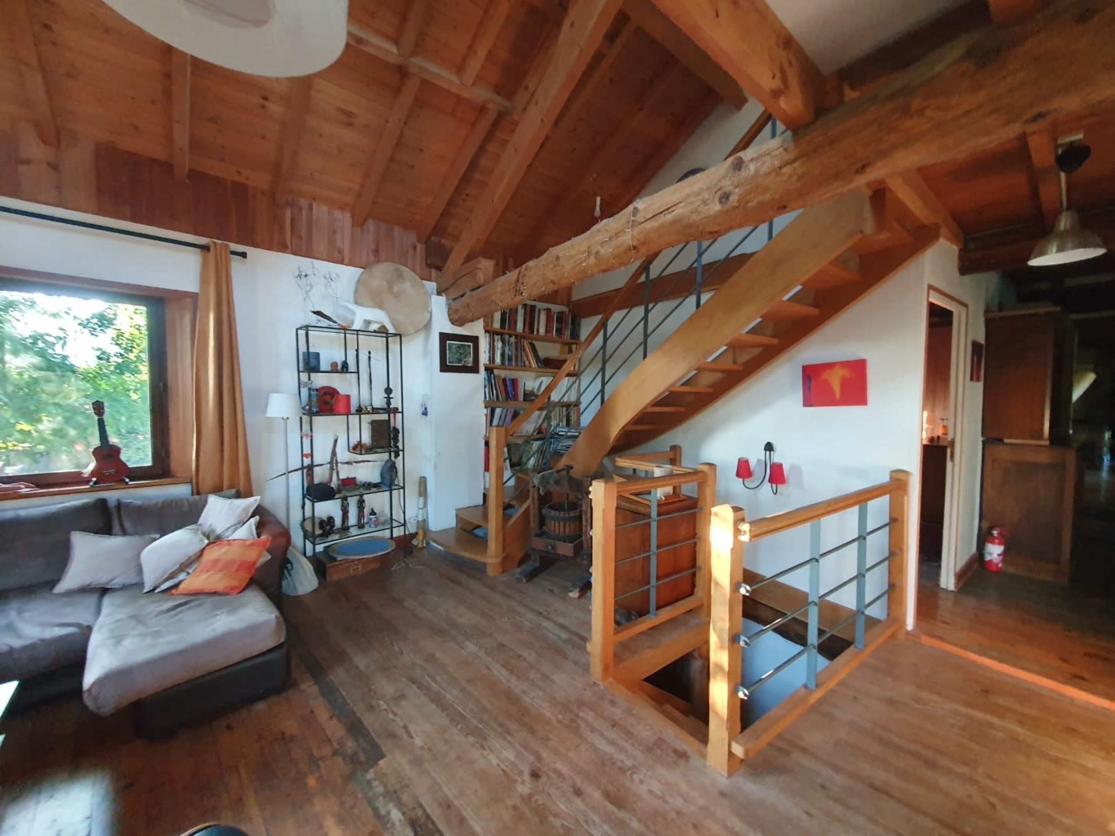 The image shows a cozy living space with wooden beams and a staircase, featuring a bookshelf, sofa, and artwork on the walls.