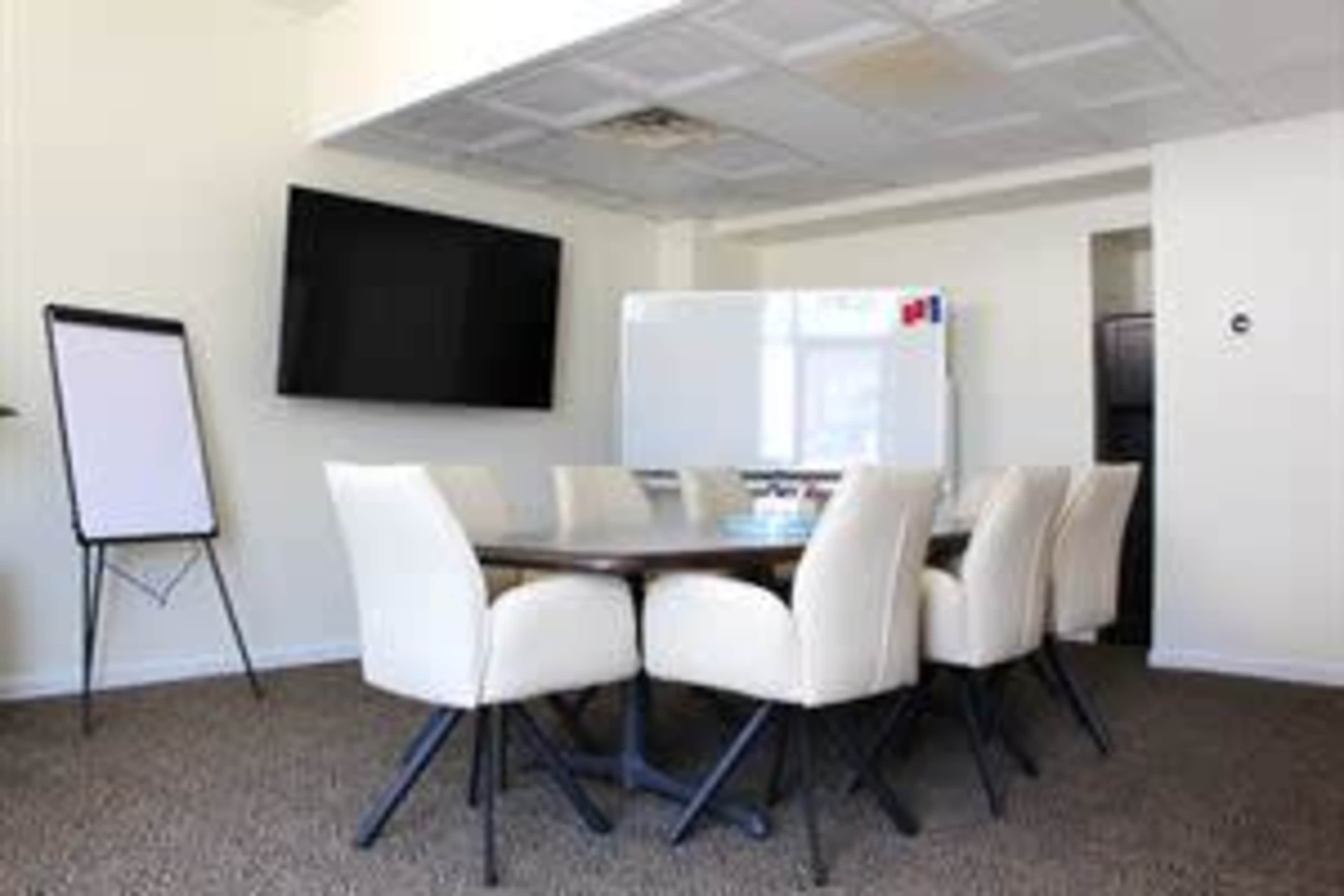 A conference room features a large table surrounded by cream-colored chairs, with a wall-mounted TV and a whiteboard nearby.