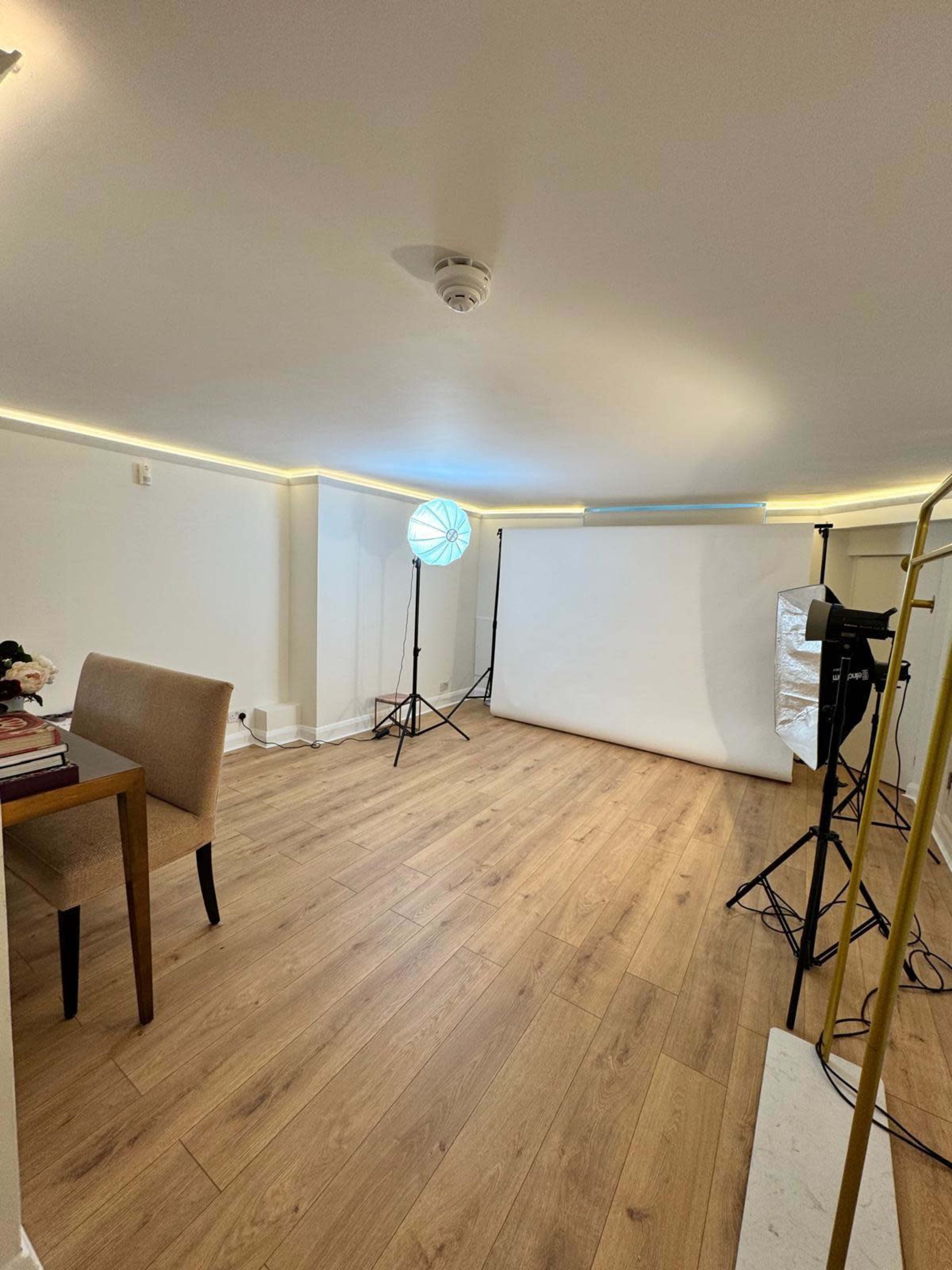 The image shows a well-lit photography studio featuring a backdrop setup, studio lights, a chair, and wooden flooring.