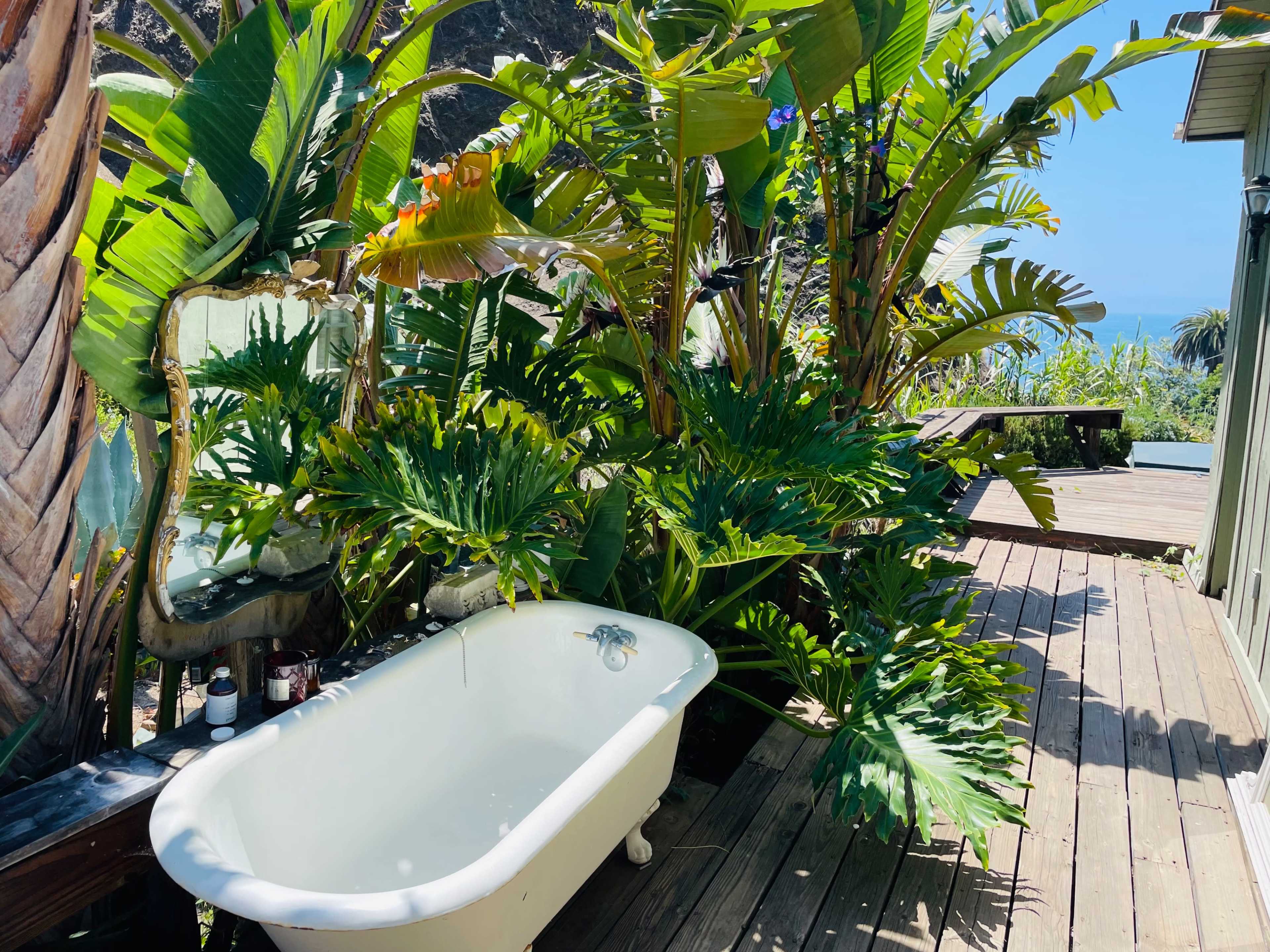 A vintage bathtub surrounded by lush tropical plants on a wooden deck with a view of the ocean.