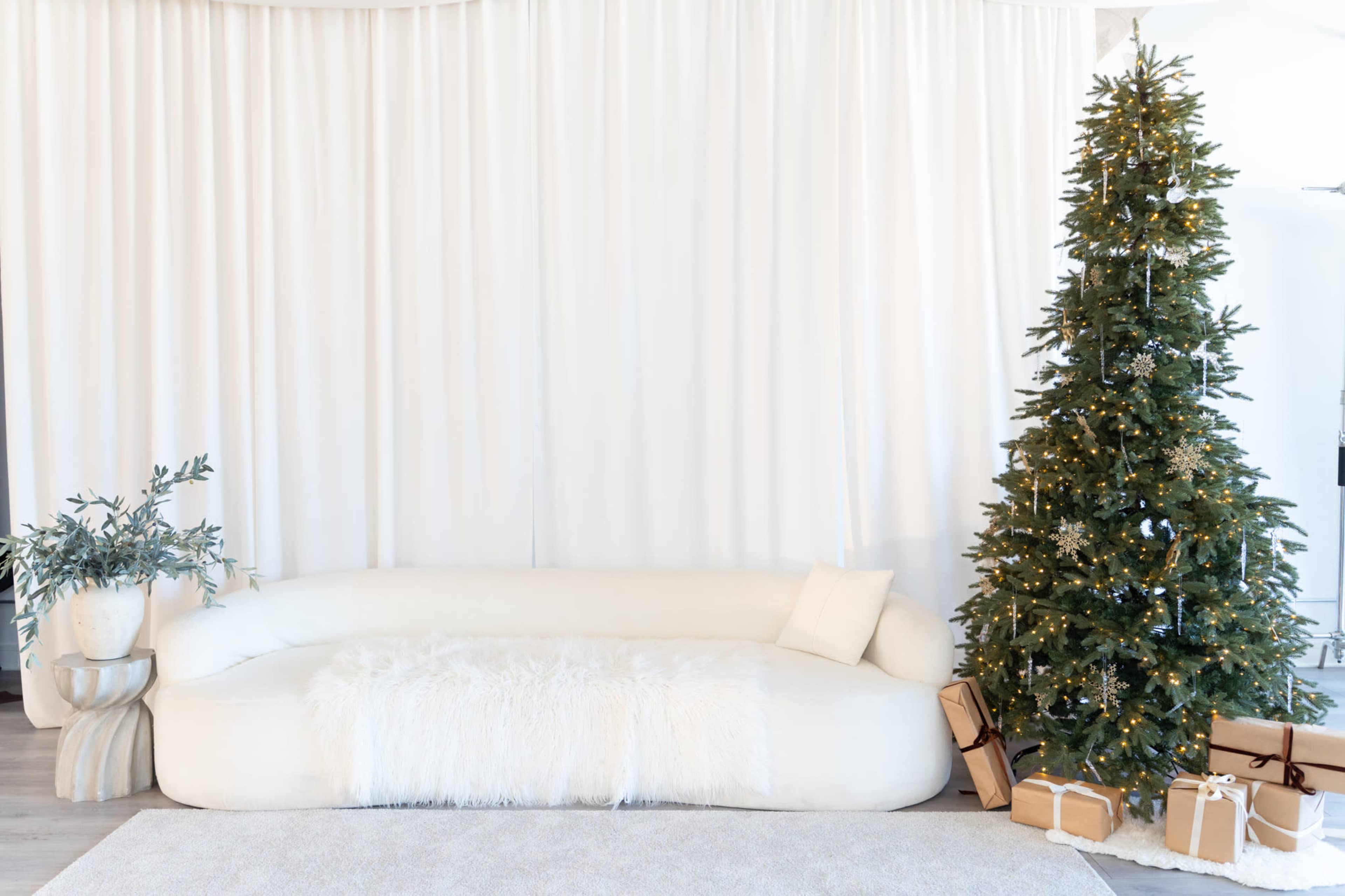 A white sectional sofa with a fur throw is positioned near a decorated Christmas tree and a potted plant in a bright, minimalist space.