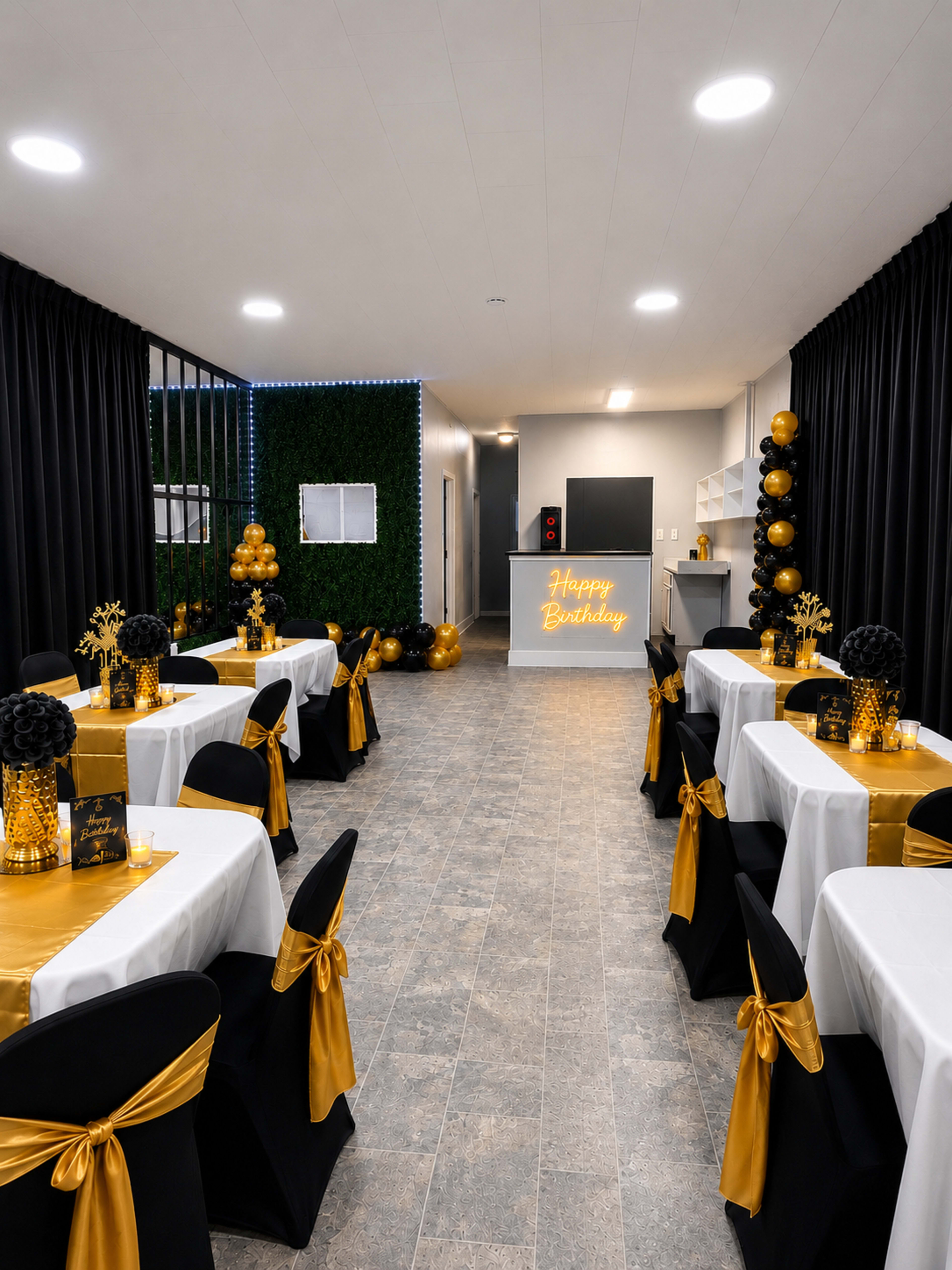 A well-decorated party venue features tables set with white cloths and gold accents, black chairs adorned with gold sashes, and a "Happy Birthday" sign illuminated at the far end.