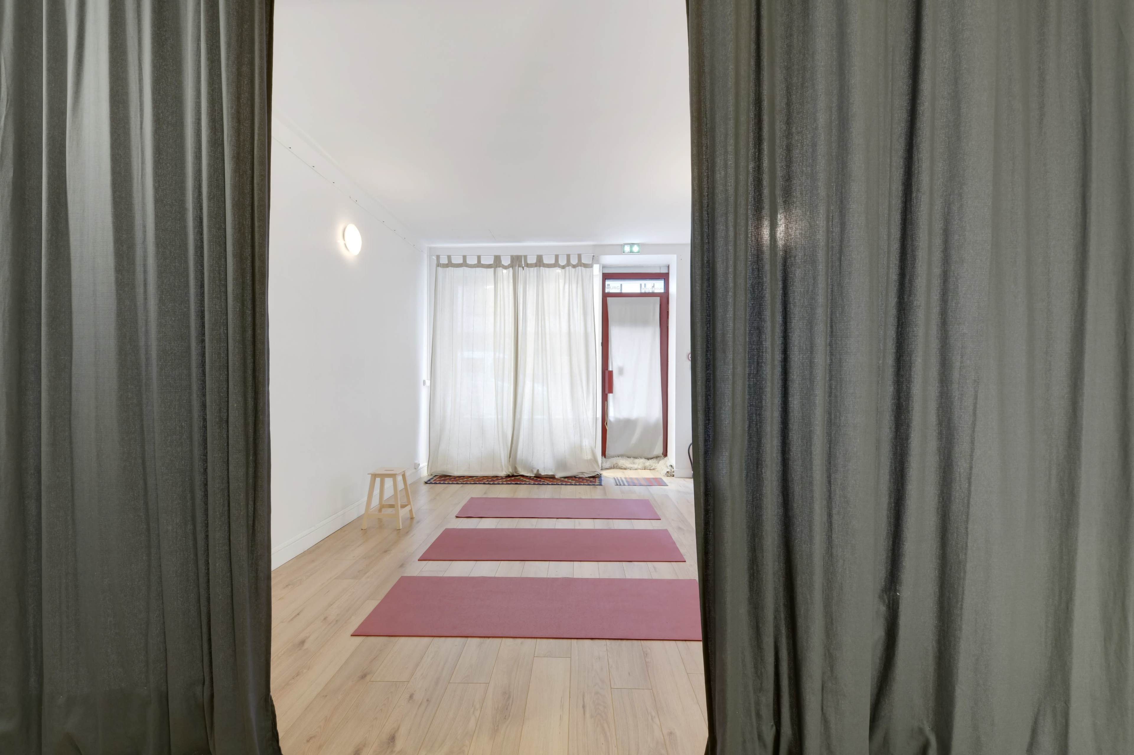 A view through curtains reveals a yoga studio with three red mats on a wooden floor and natural light coming from a window.