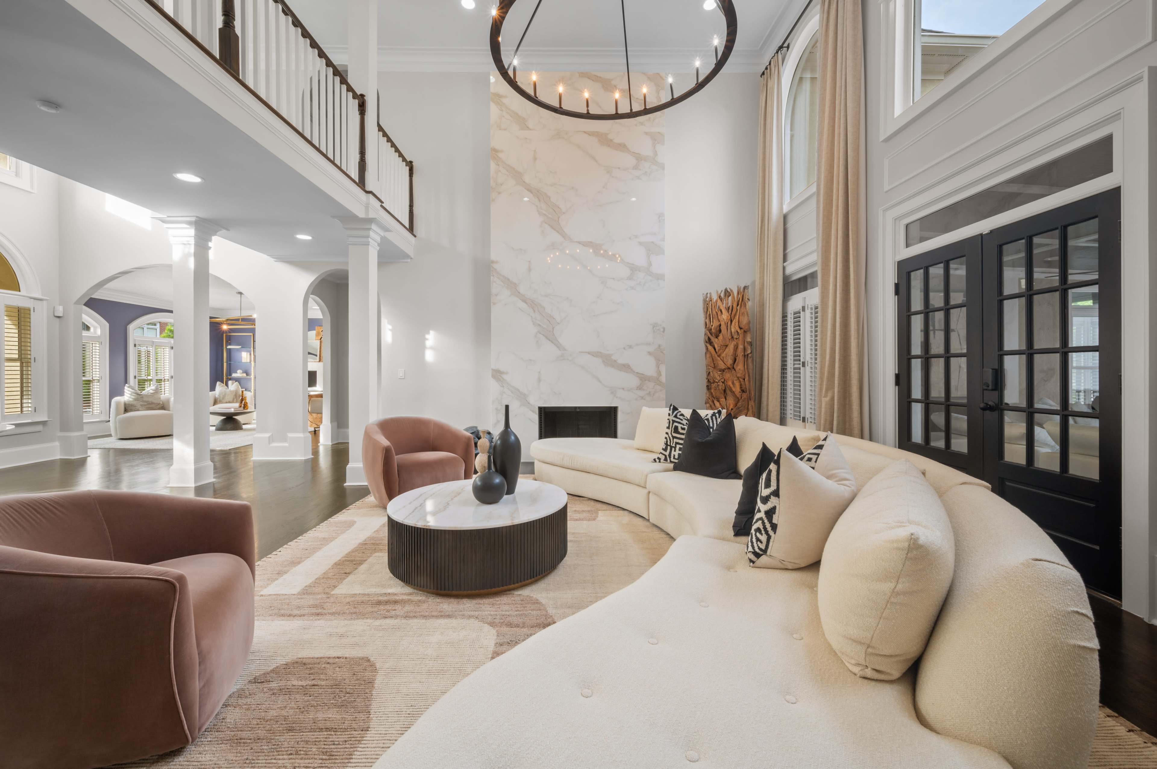 A large, modern living room features a curved sofa, a round coffee table, and a light-colored marble accent wall under a circular chandelier.