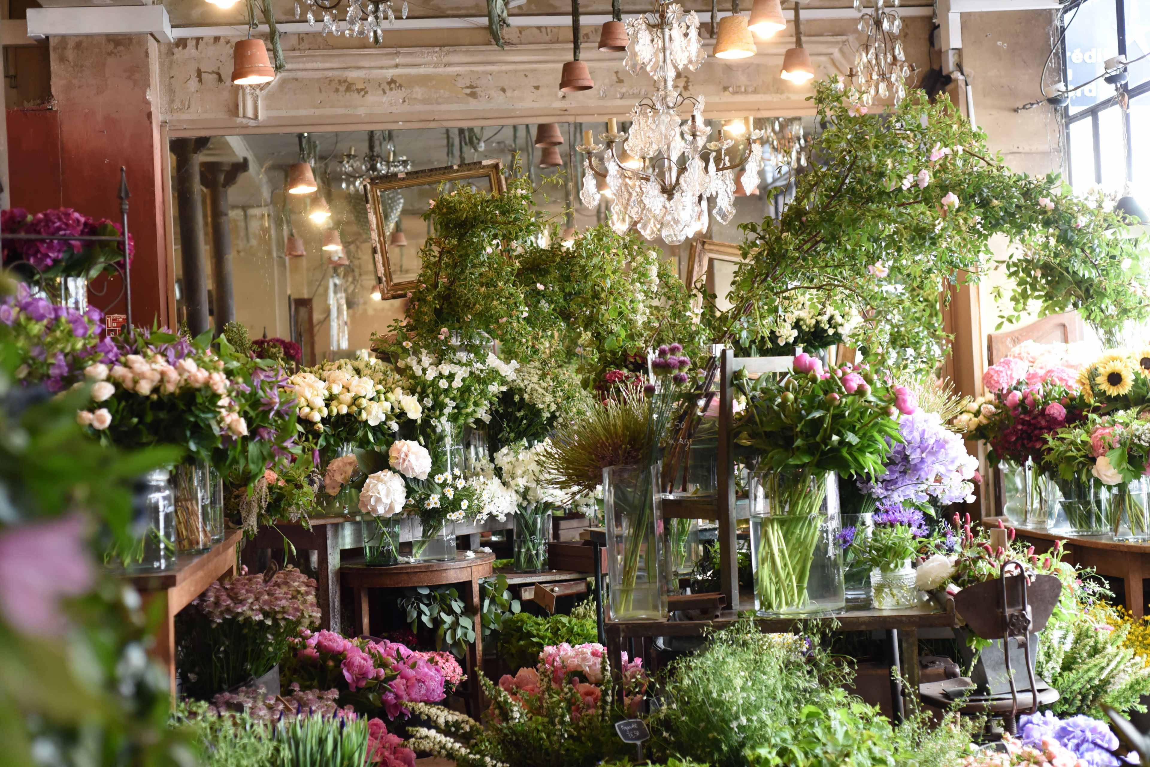A floral shop filled with a variety of colorful flowers arranged in glass vases on wooden tables, under a chandelier and surrounded by mirrors.