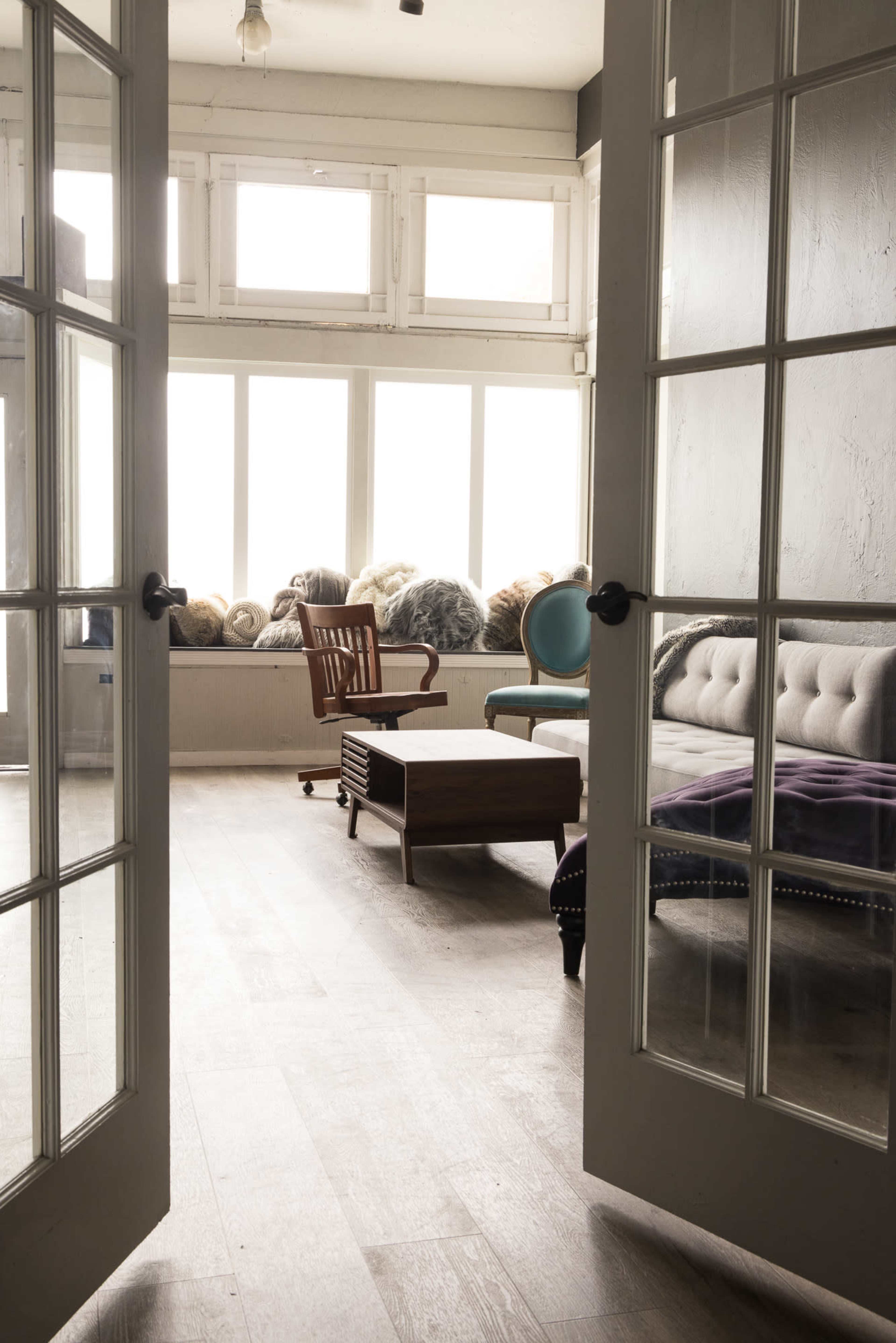 A brightly lit room features two sets of open French doors, with a tufted sofa, a wooden chair, and a coffee table visible inside.