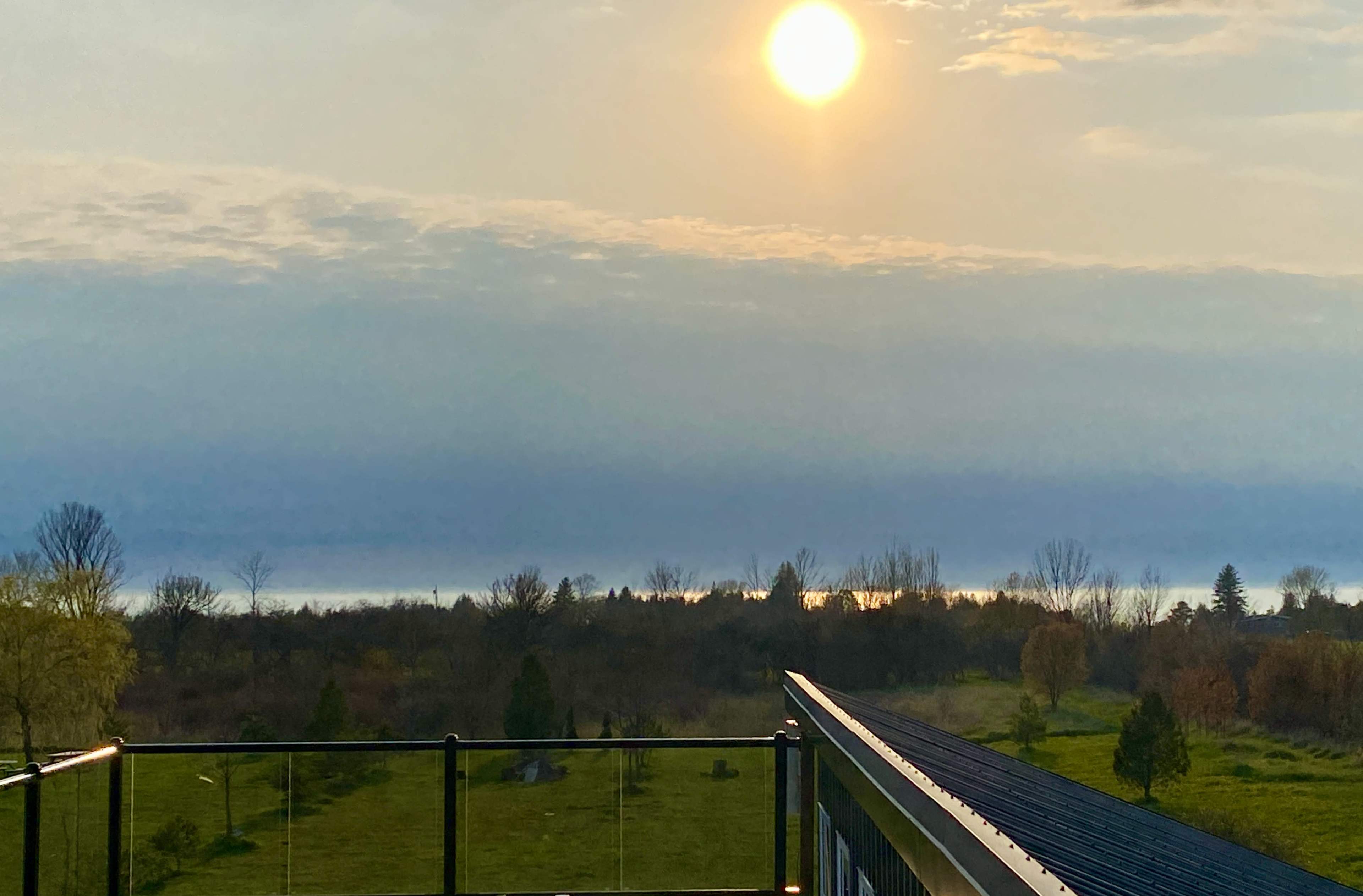 A sunrise over a tranquil landscape, with a railing in the foreground and a reflection of the sun on the water in the distance.