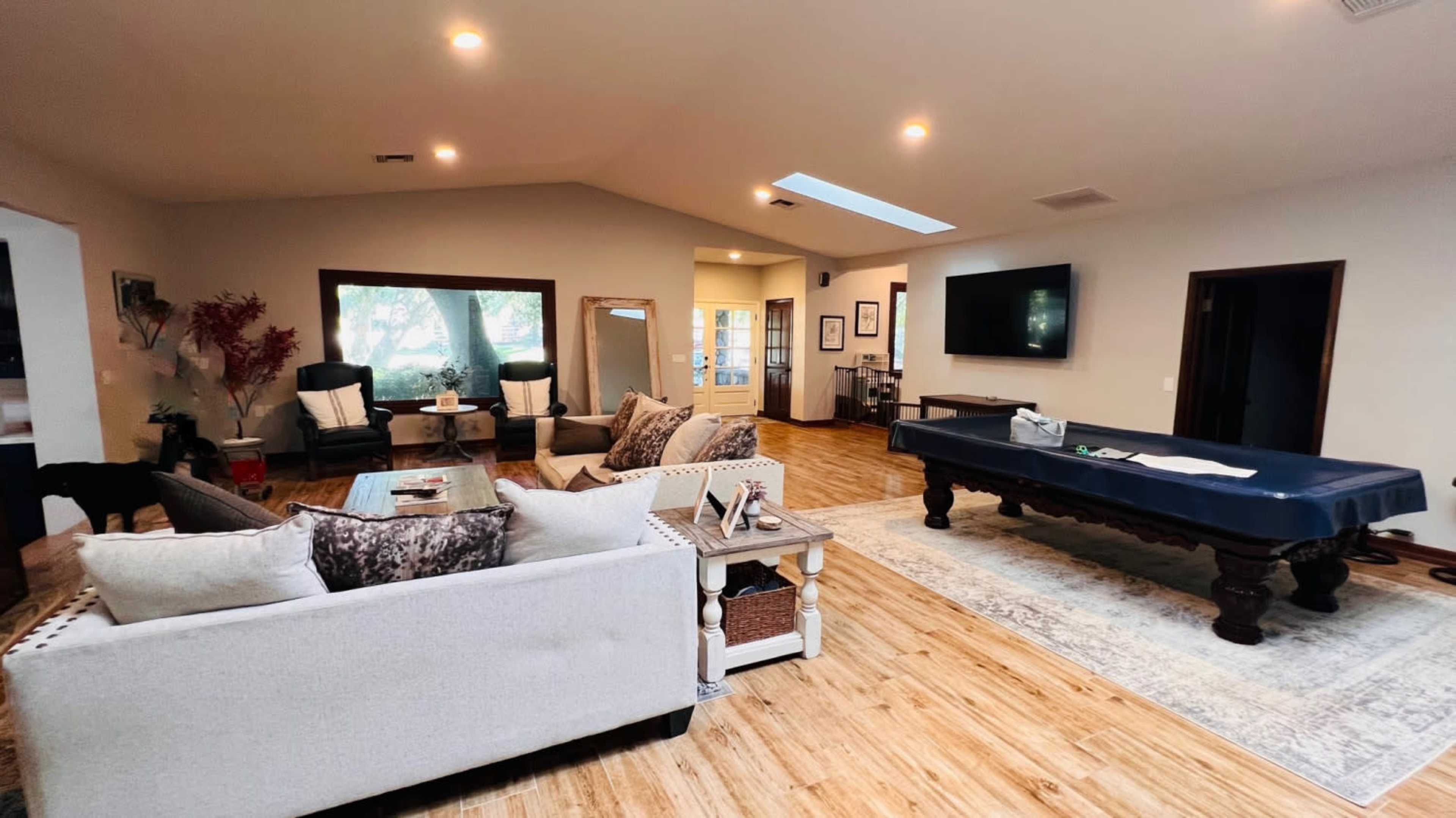 A spacious living room with a pool table, two armchairs, and a sofa arranged around a coffee table, with large windows allowing natural light to enter.