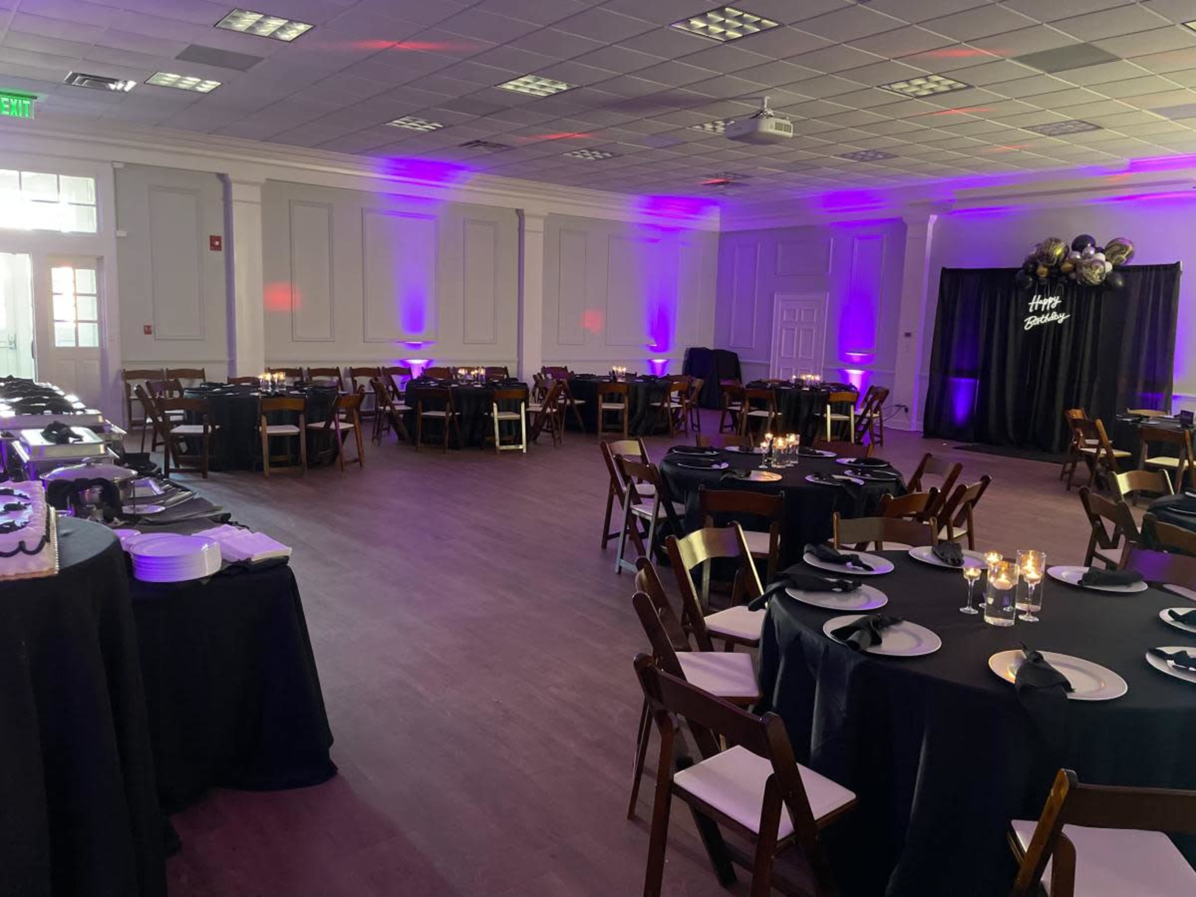 The interior of a banquet hall features neatly arranged round tables with black tablecloths, wooden chairs, and purple lighting, set up for an event.