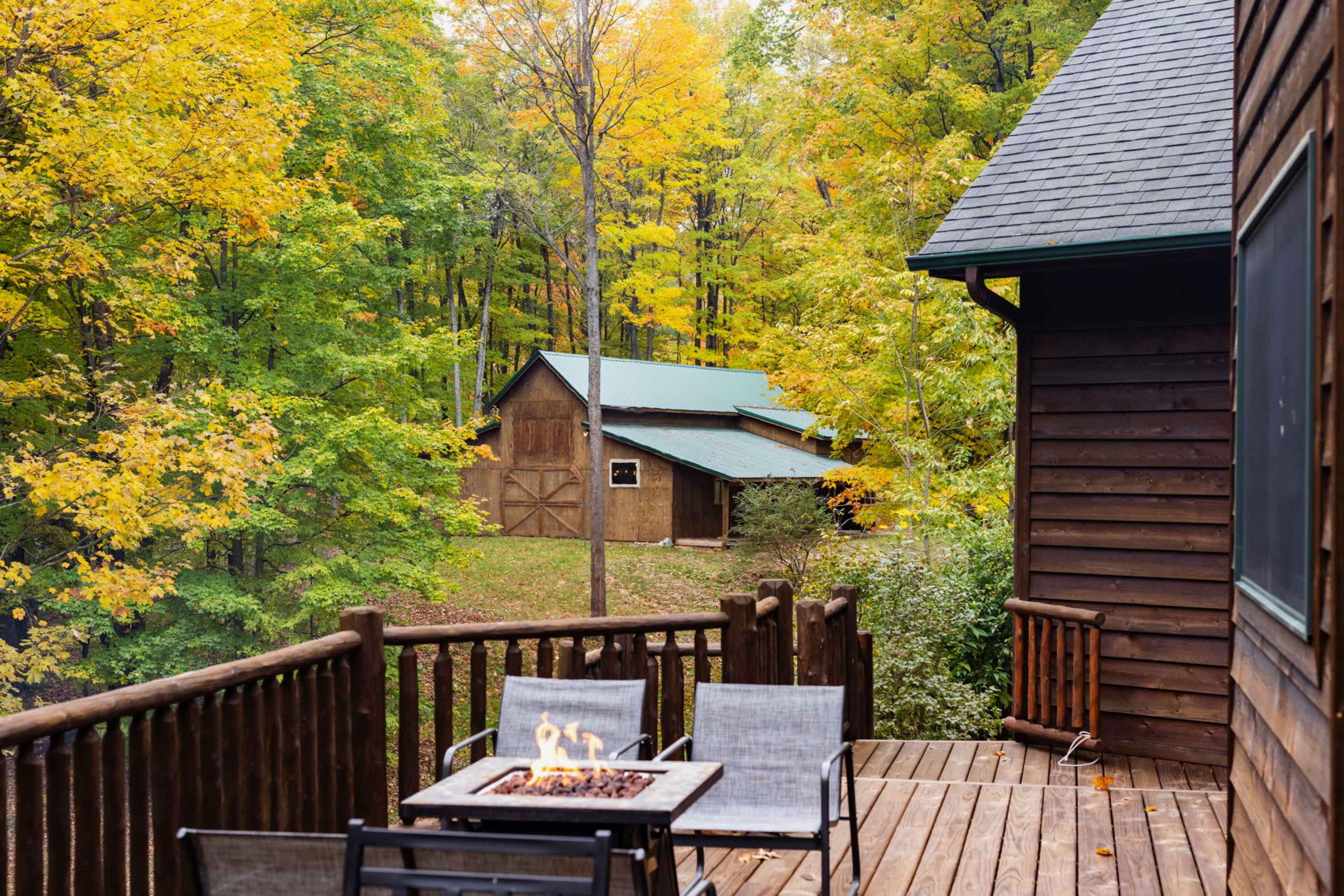 A wooden deck with a fire pit faces a cozy cabin amidst trees displaying autumn foliage.