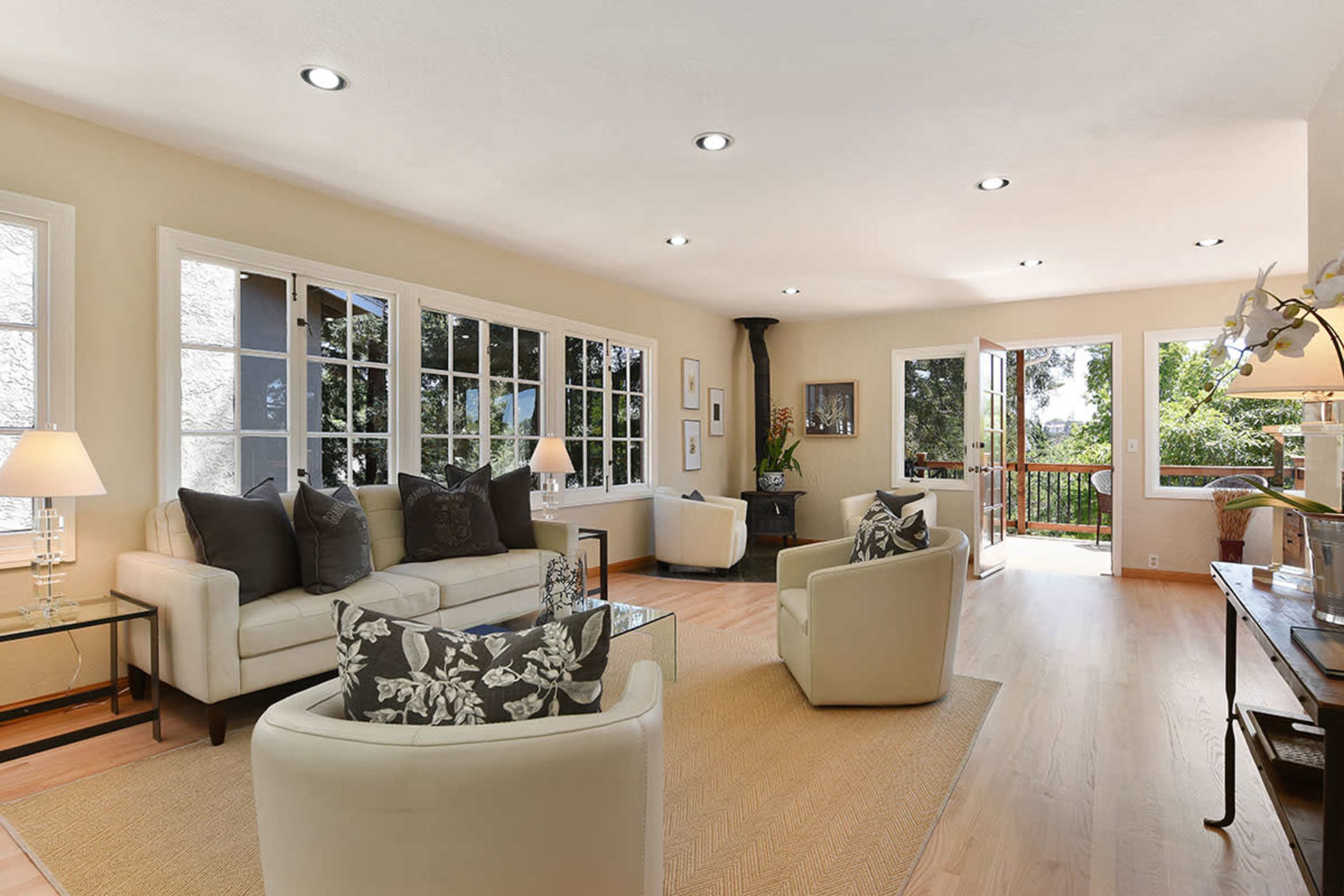 The bright, open living room features two light-colored sofas, a coffee table, and large windows that lead to a balcony with outdoor views.