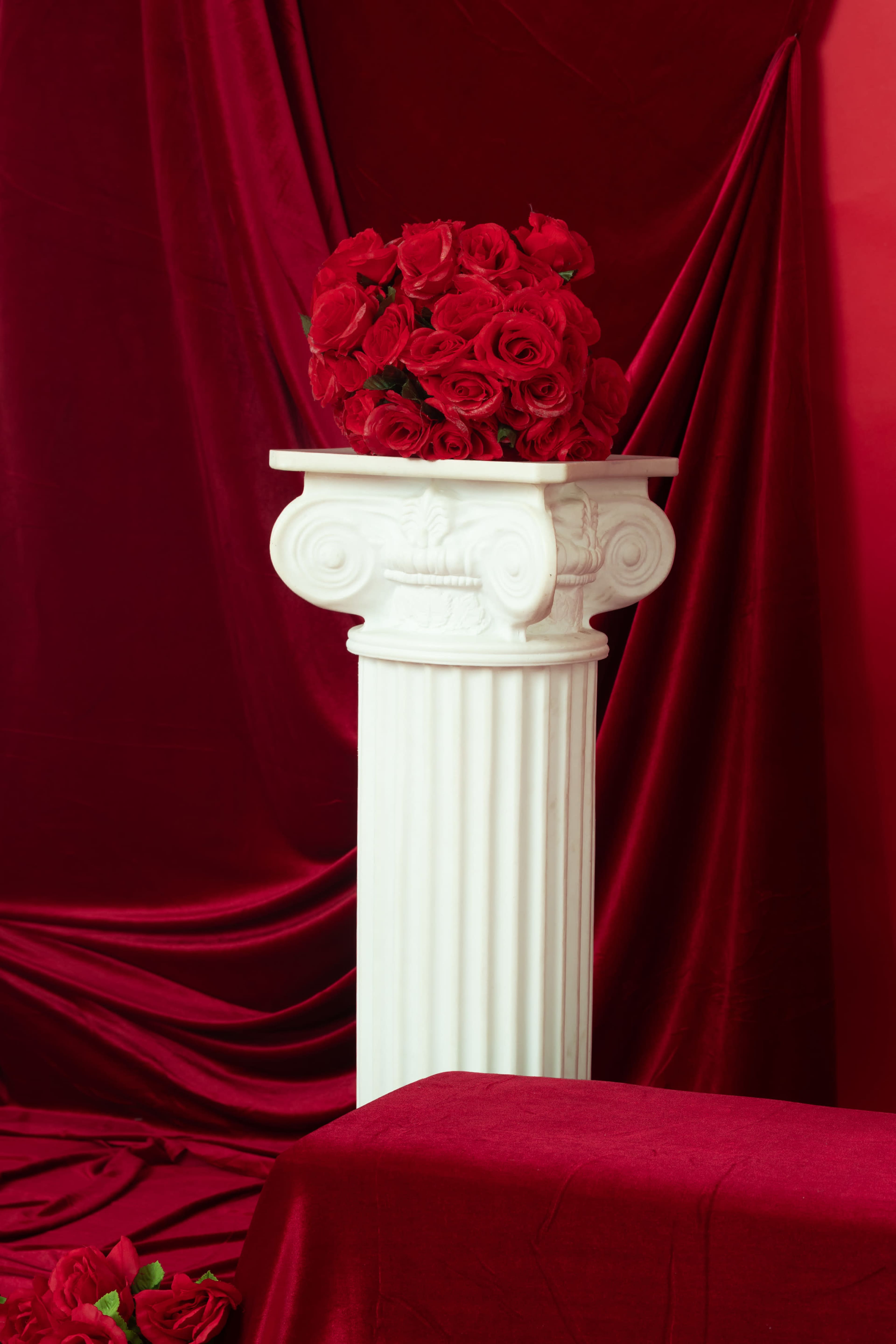 A white marble column supports a bouquet of red roses, set against a backdrop of rich red fabric.