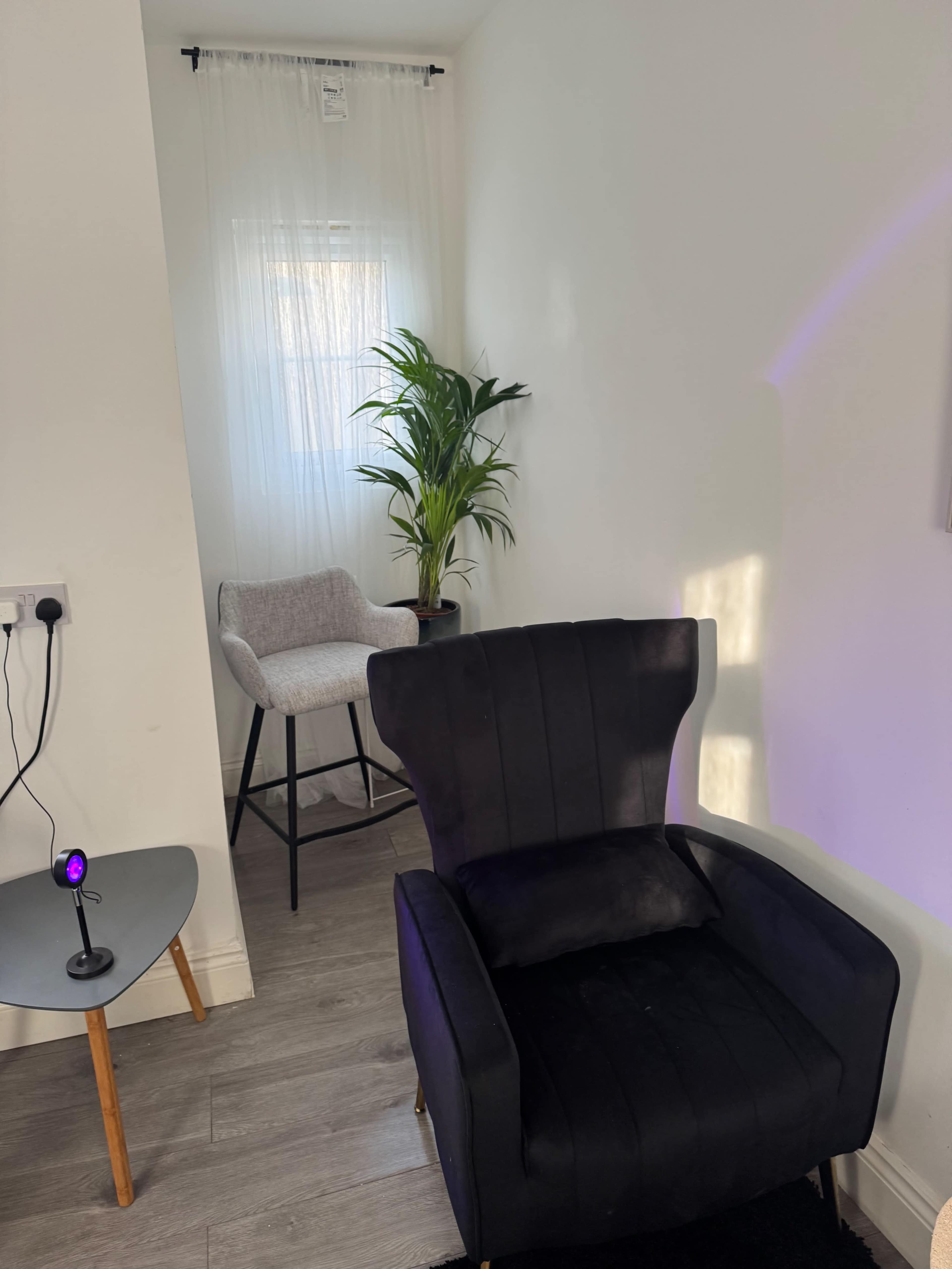 The image shows a small room with a black armchair, a light gray bar stool, and a potted plant by a window with sheer curtains.