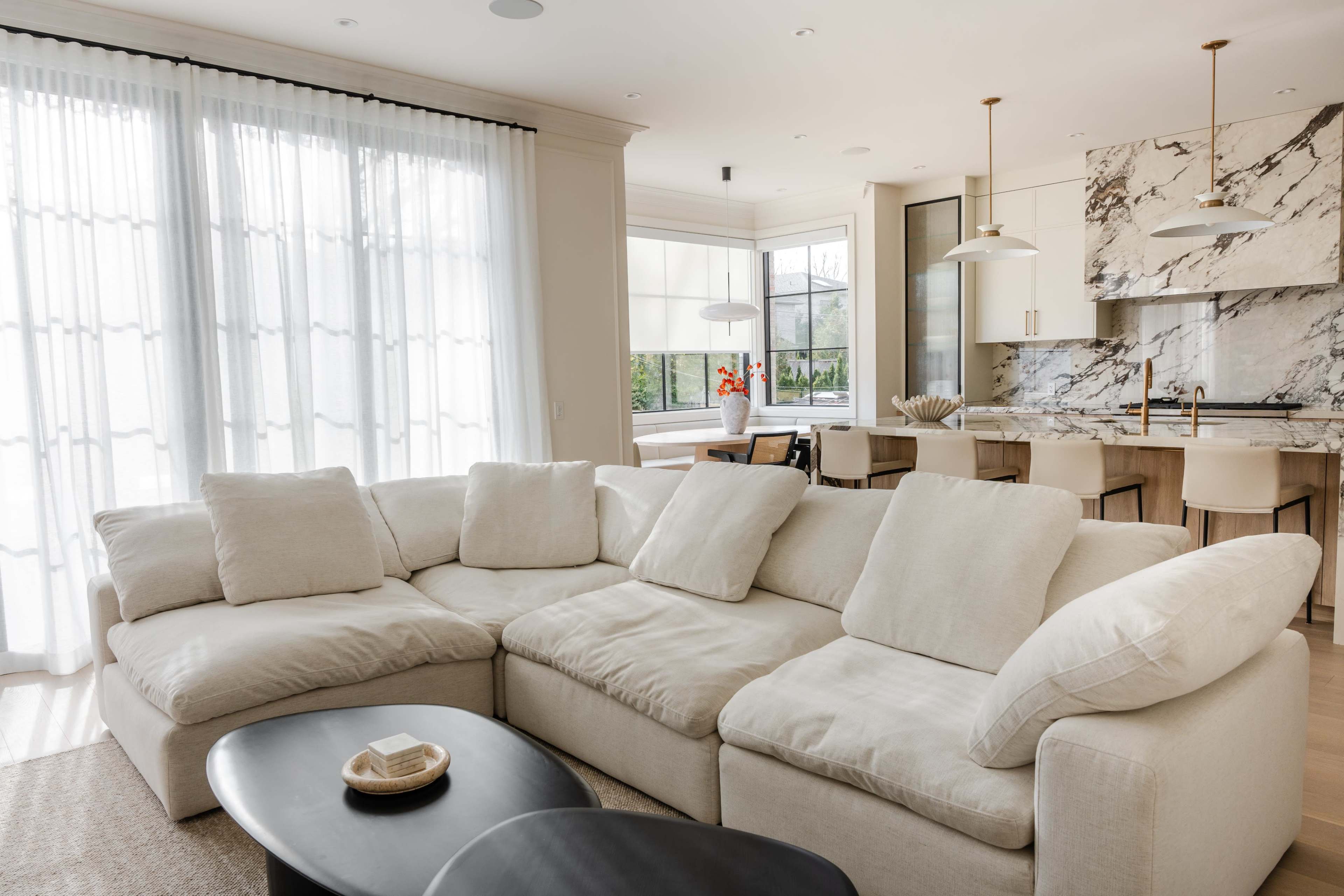 A spacious living area features a large sectional sofa, a round black coffee table, and a marble kitchen in the background with large windows providing natural light.