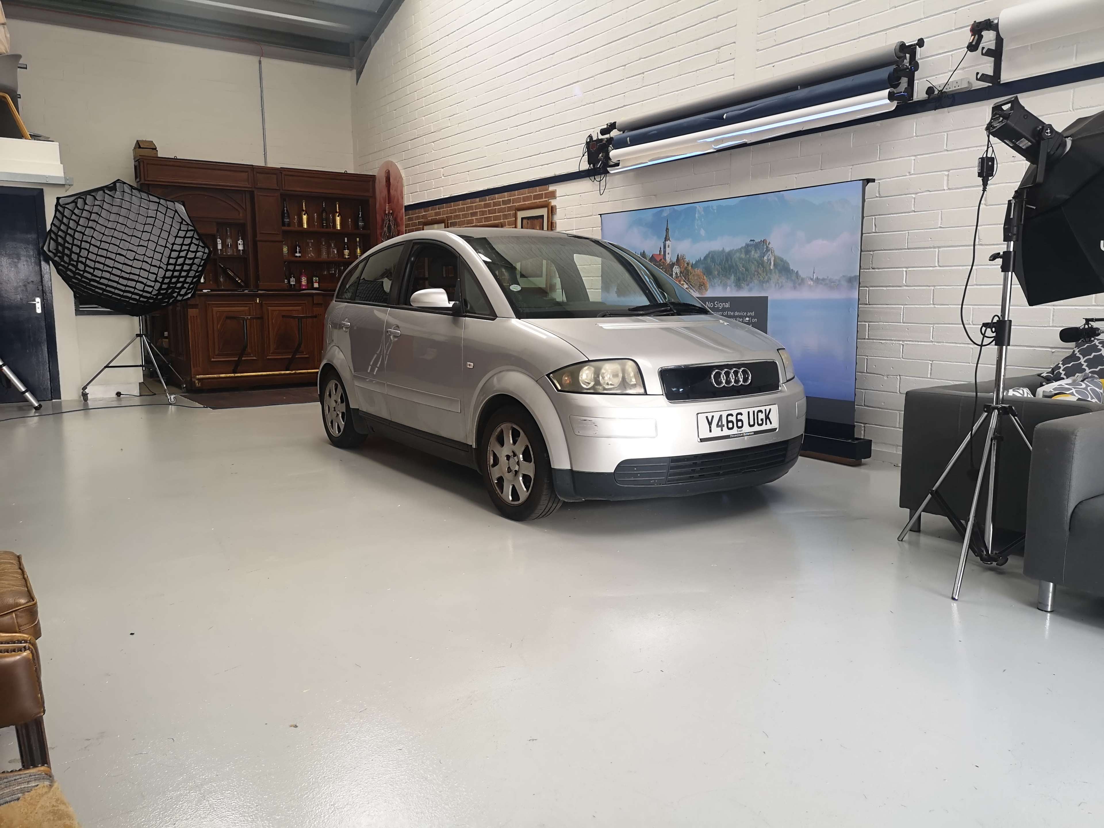 A silver Audi car is parked in a spacious, well-lit room with a wooden cabinet, a backdrop featuring a scenic view, and photography equipment.